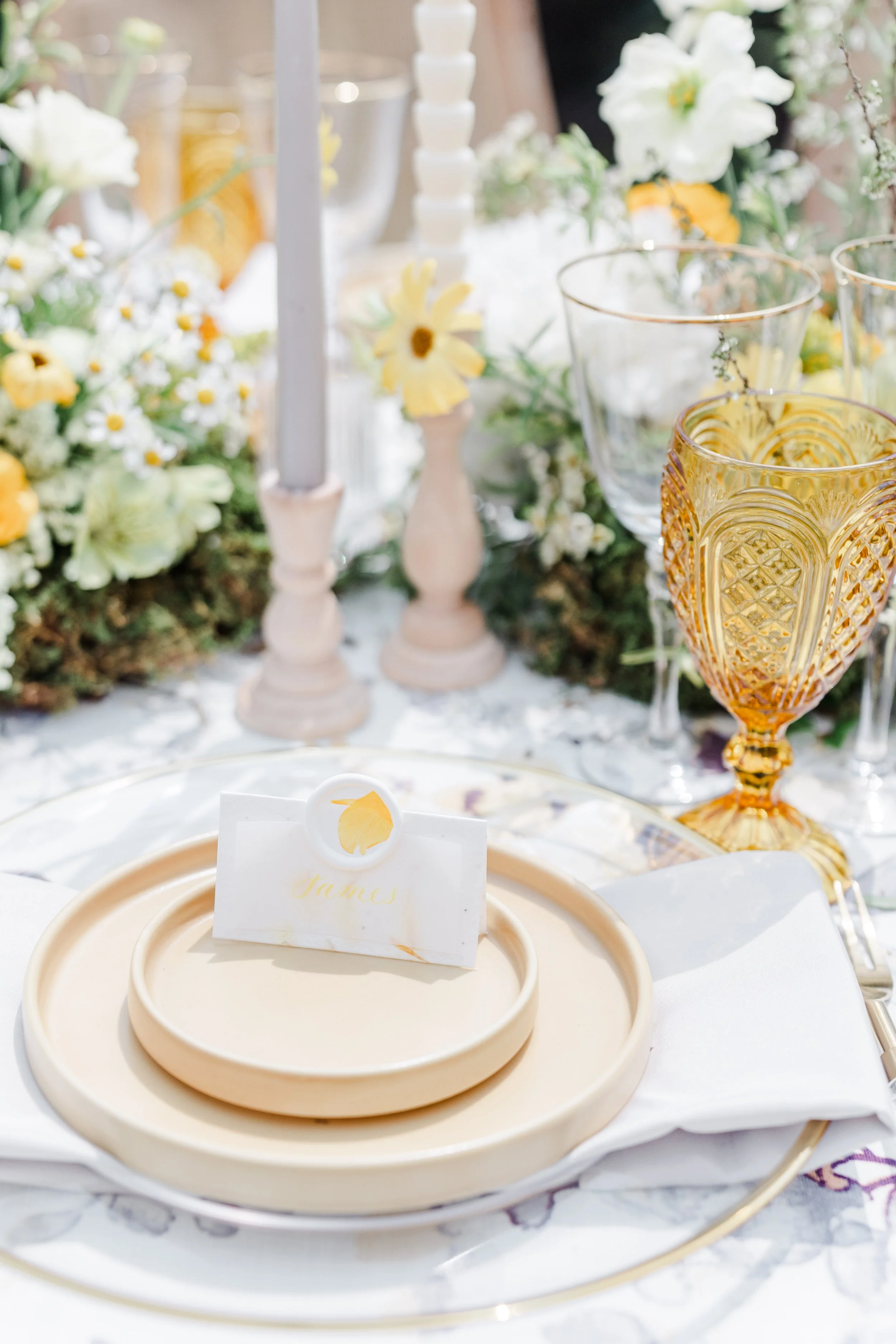 Elegant table setting with beige and gold plates, white napkin, and a name card that reads 'James', surrounded by flowers, candles, and glassware.