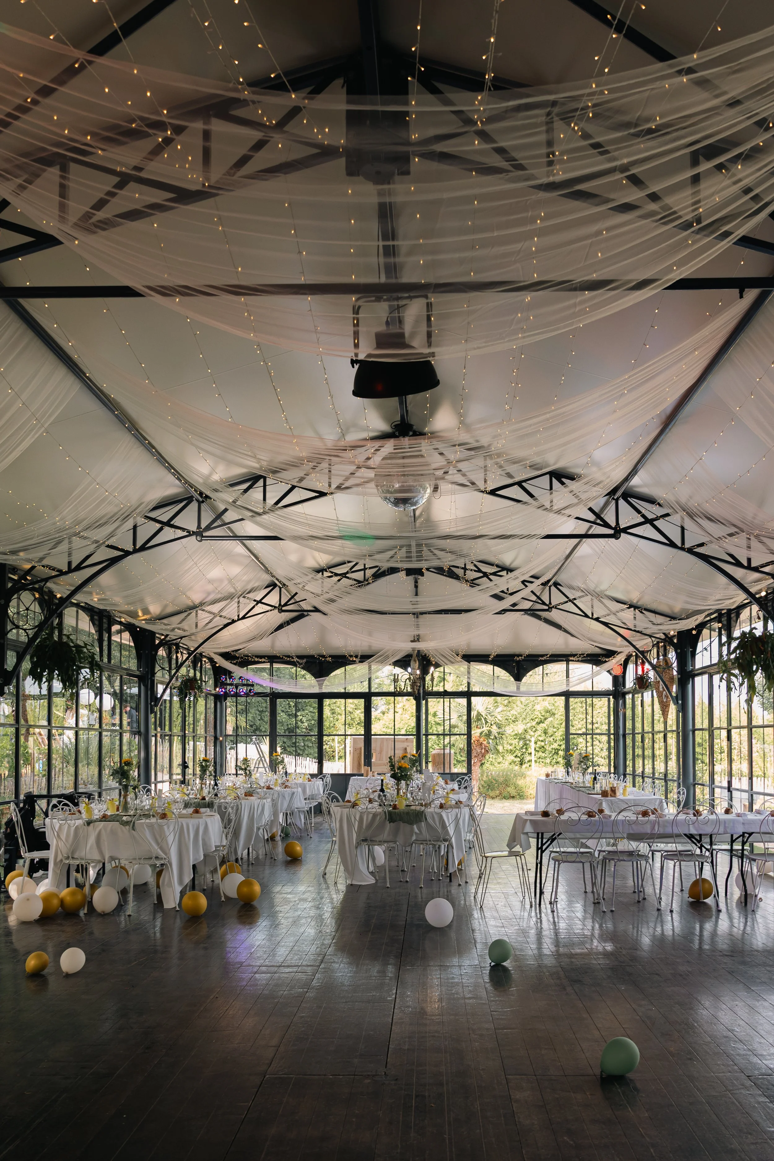 Salle de réception décorée avec des tables habillées de nappes blanches, des ballons dorés et blancs sur le sol, et un plafond décoré de rideaux transparents et de guirlandes de lumières, avec de grandes fenêtres laissant entrer la lumière naturelle.