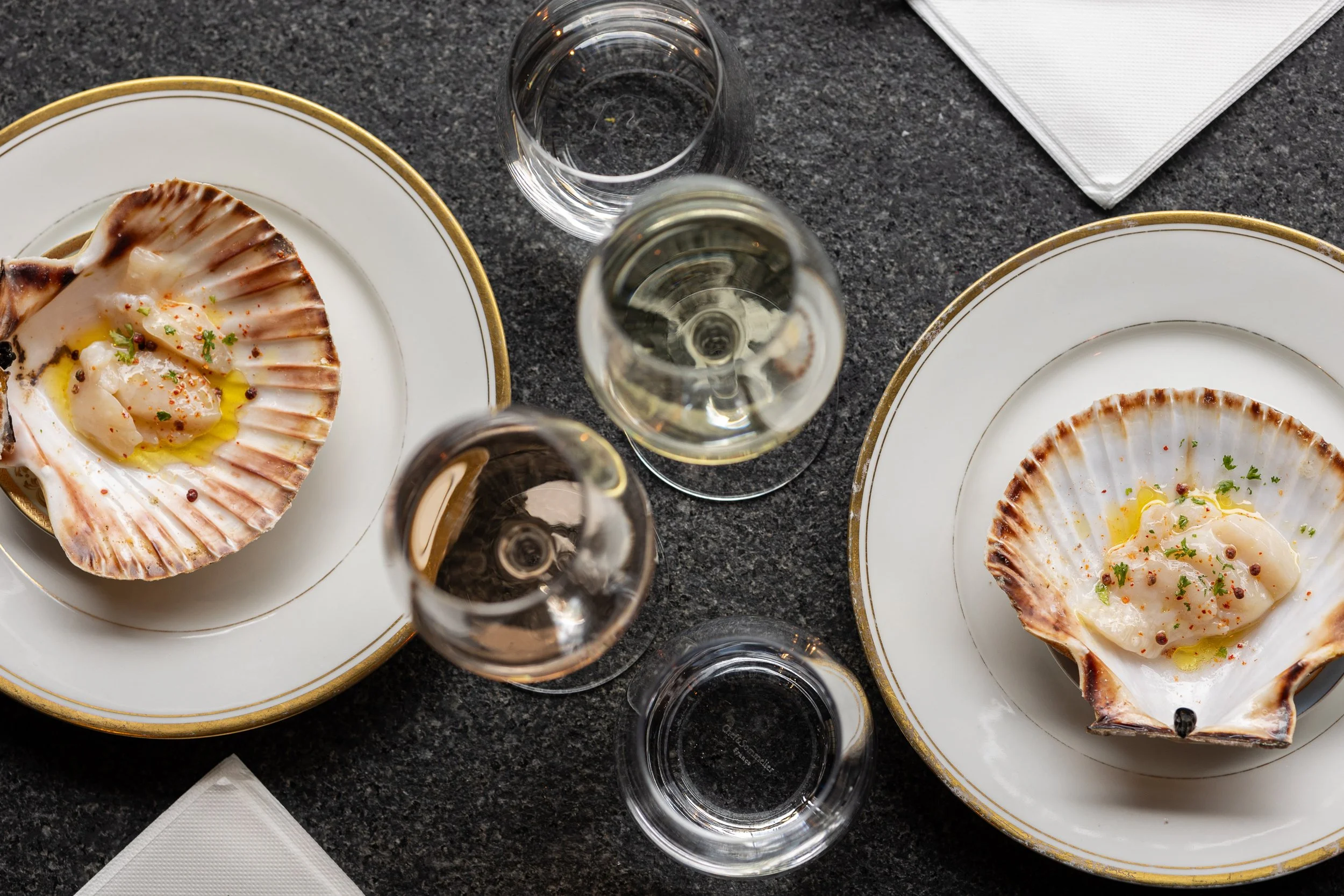 Deux coquilles Saint-Jacques garnies, accompagnées de verres de vin blanc, sur une table en pierre