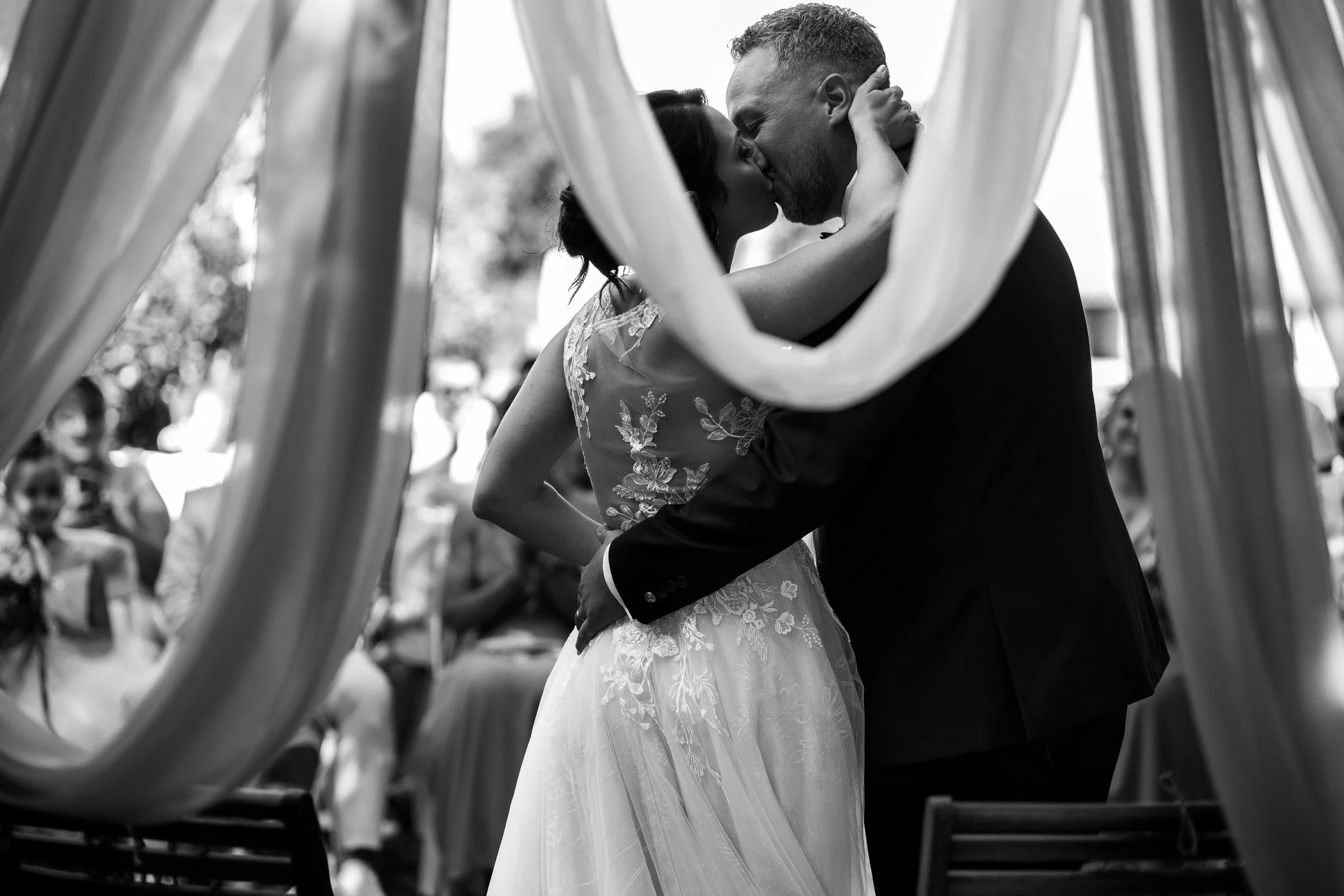 Un couple de mariés s'embrassant lors de leur mariage, vus à travers des draperies, en noir et blanc.