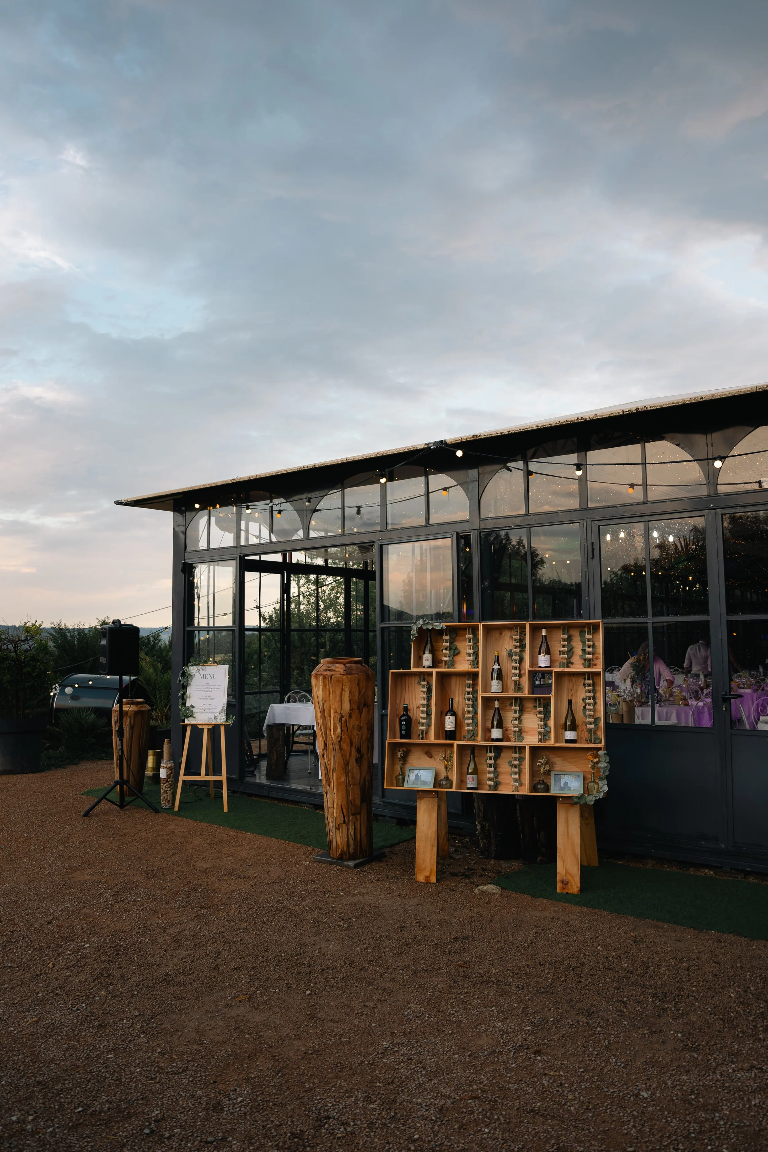 Un bâtiment en verre avec des tables décorées pour un événement, des bouteilles de vin sur une étagère en bois, et un ciel nuageux en arrière-plan.