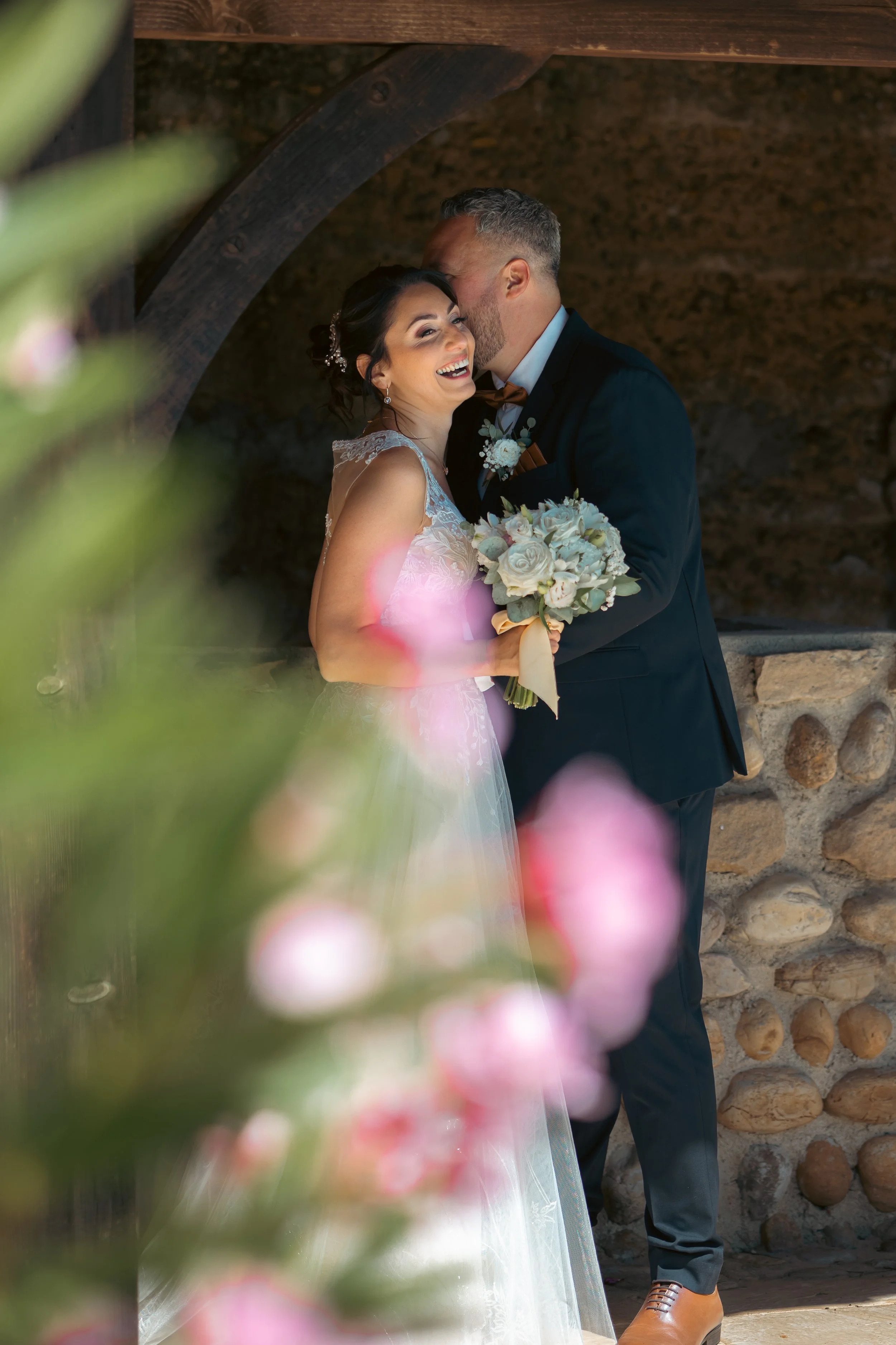 Un couple de mariés lors de leur mariage, la femme tenant un bouquet de fleurs blanches, sourit et rit pendant que l'homme lui embrasse le front.