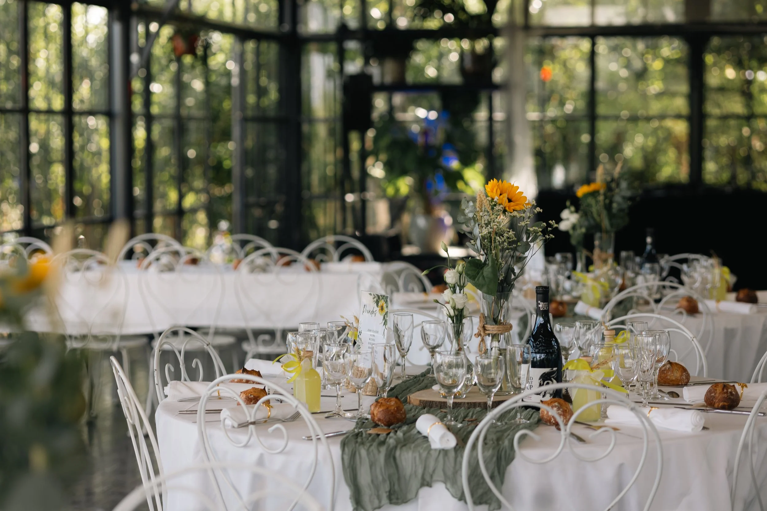 Table décorée avec des fleurs, des bouteilles de champagne, des verres, et des pains, dans un cadre intérieur lumineux avec beaucoup de fenêtres laissant passer la lumière naturelle.