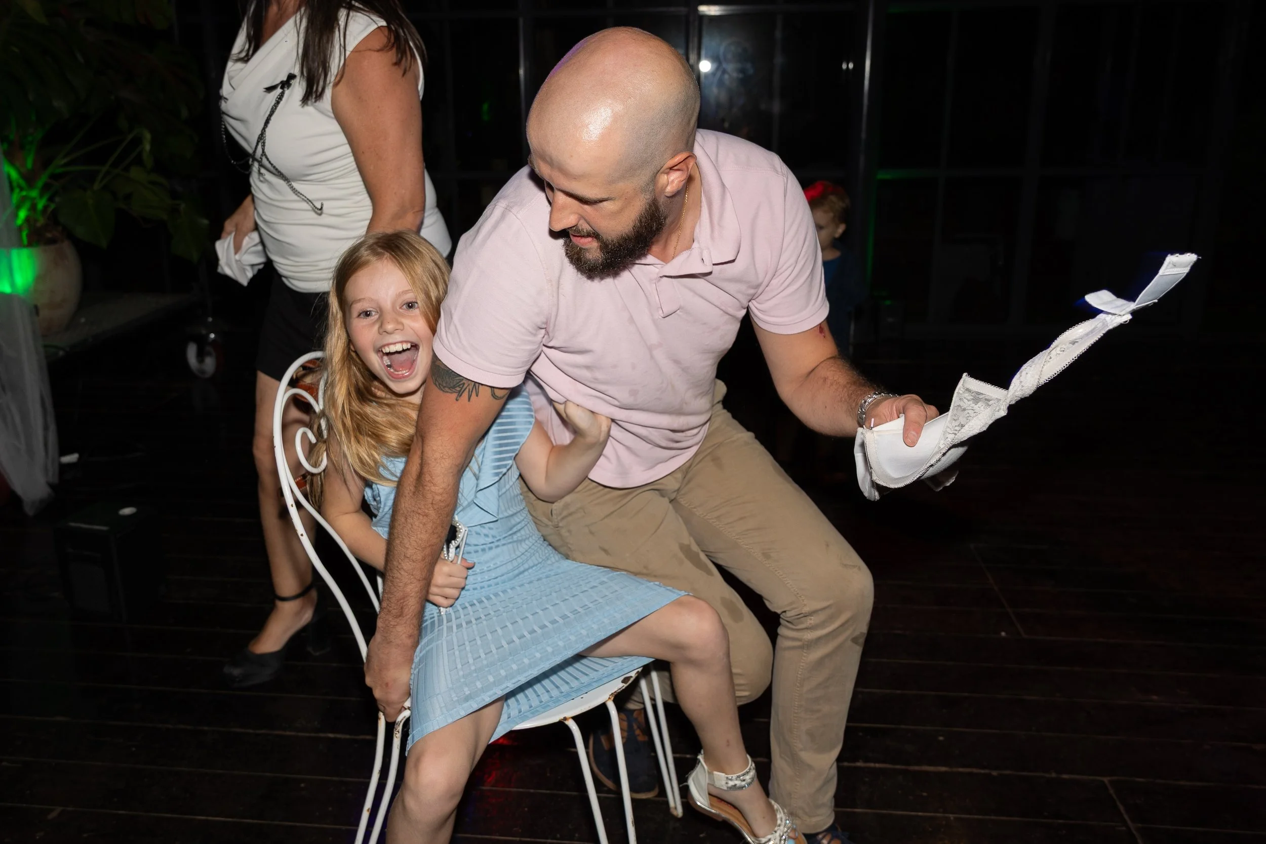 Un homme et une jeune fille s'amusent à une fête, avec la jeune fille assise sur une chaise en métal blanche, riant joyeusement, tandis que l'homme semble la faire danser ou jouer. La scène se passe dans une pièce sombre avec un fond noir et des écla
