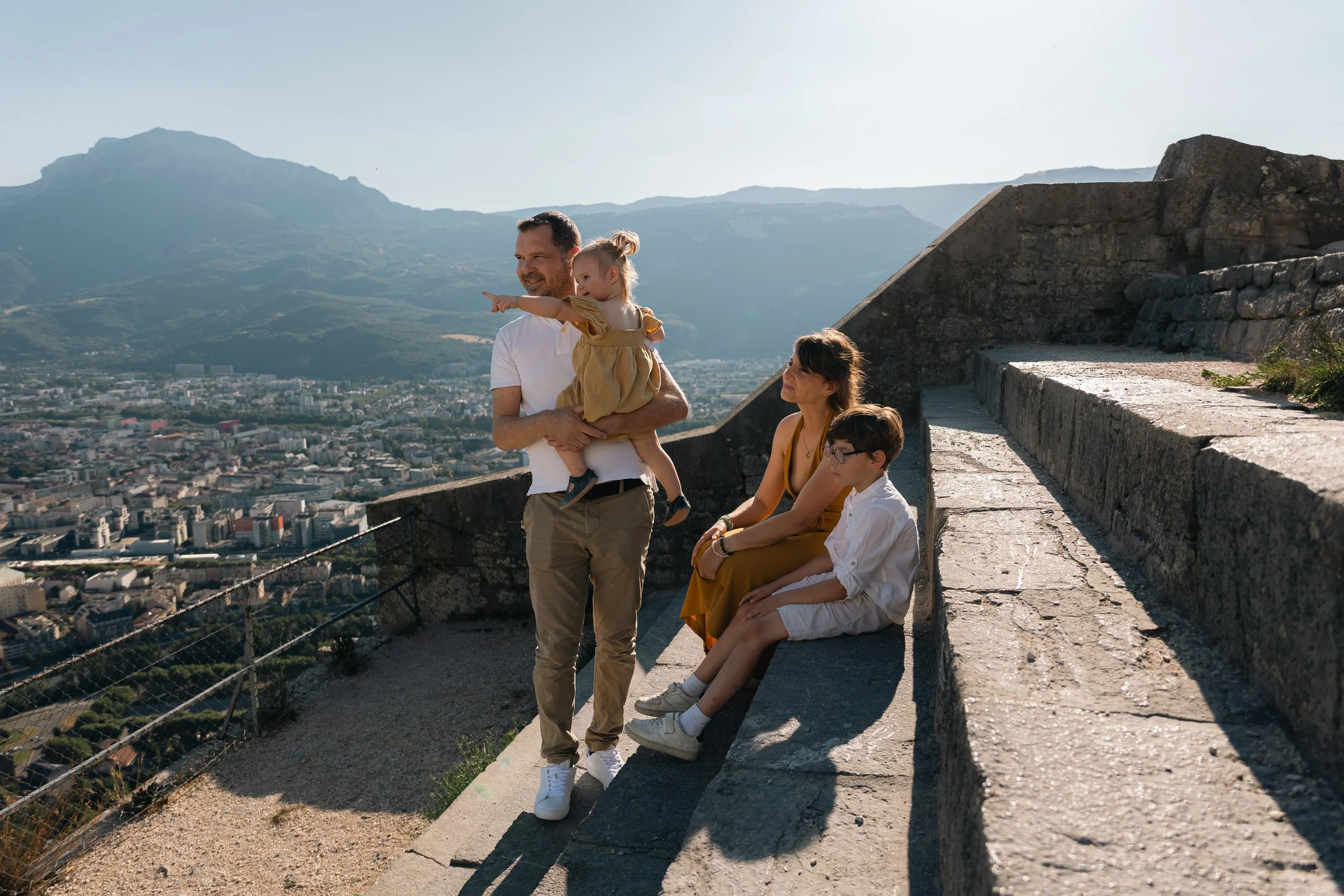 Un groupe familial de quatre personnes profite d'une vue panoramique en haut d'une colline ou d'une structure historique. Le père, portant une chemise blanche, porte une jeune fille en robe beige qui pointe du doigt vers l'horizon. La mère, en robe j