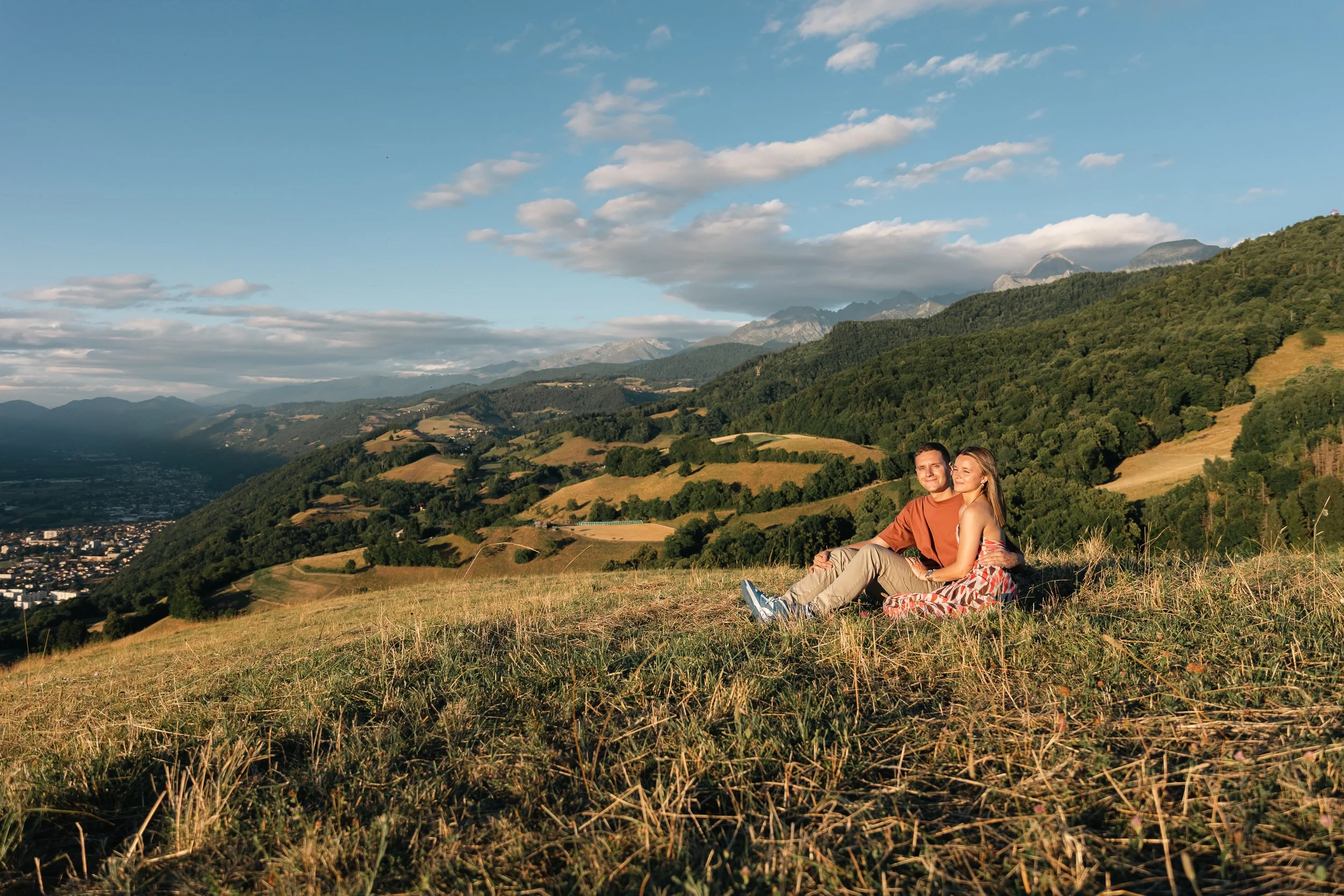 Un couple assis dans l'herbe sur une colline avec un paysage de montagnes, de vallées et un ciel bleu avec des nuages en arrière-plan.