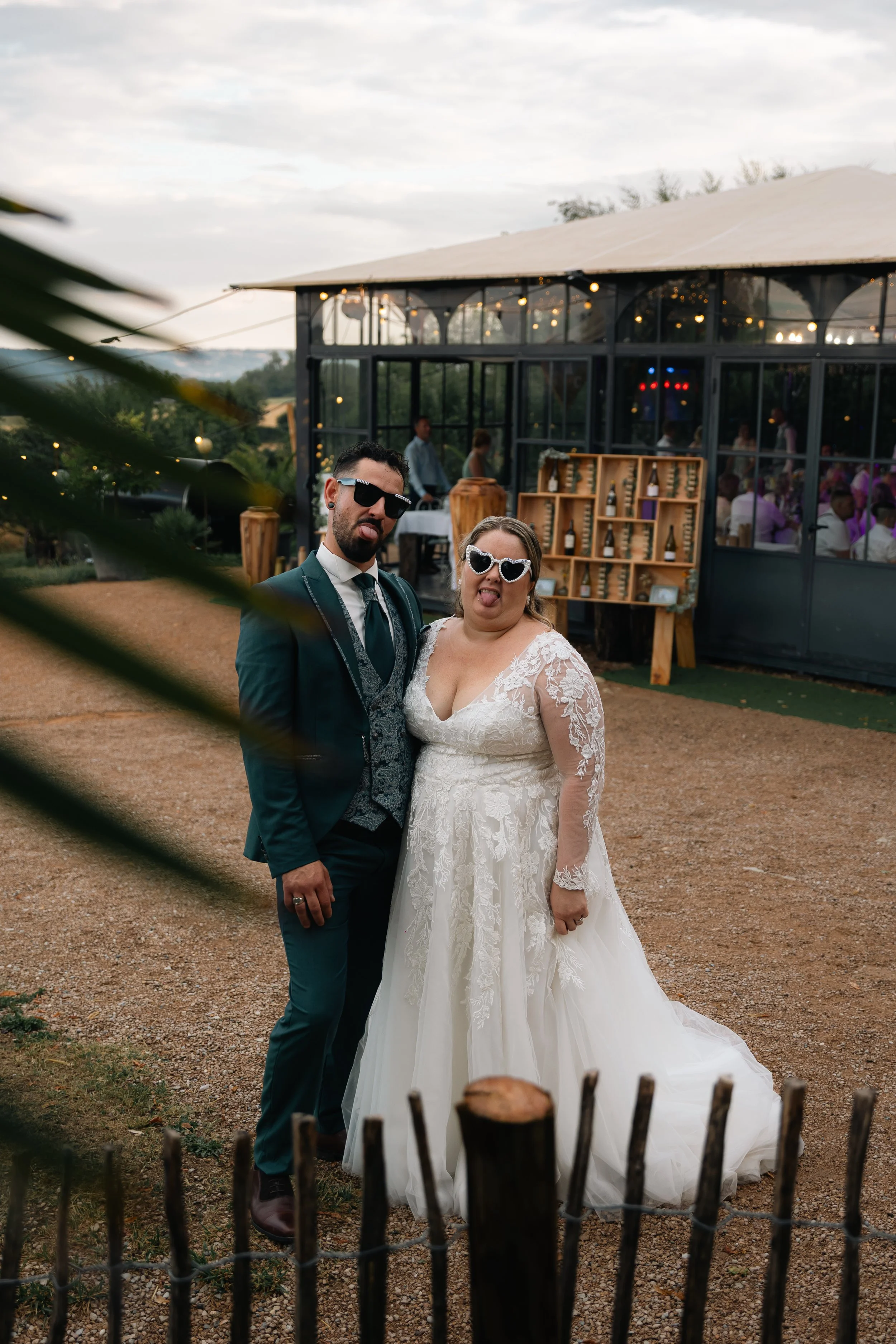 Un couple habillé en mariage, faisant la fête, portant des lunettes de soleil, dans un cadre extérieur lors d'une réception en soirée.