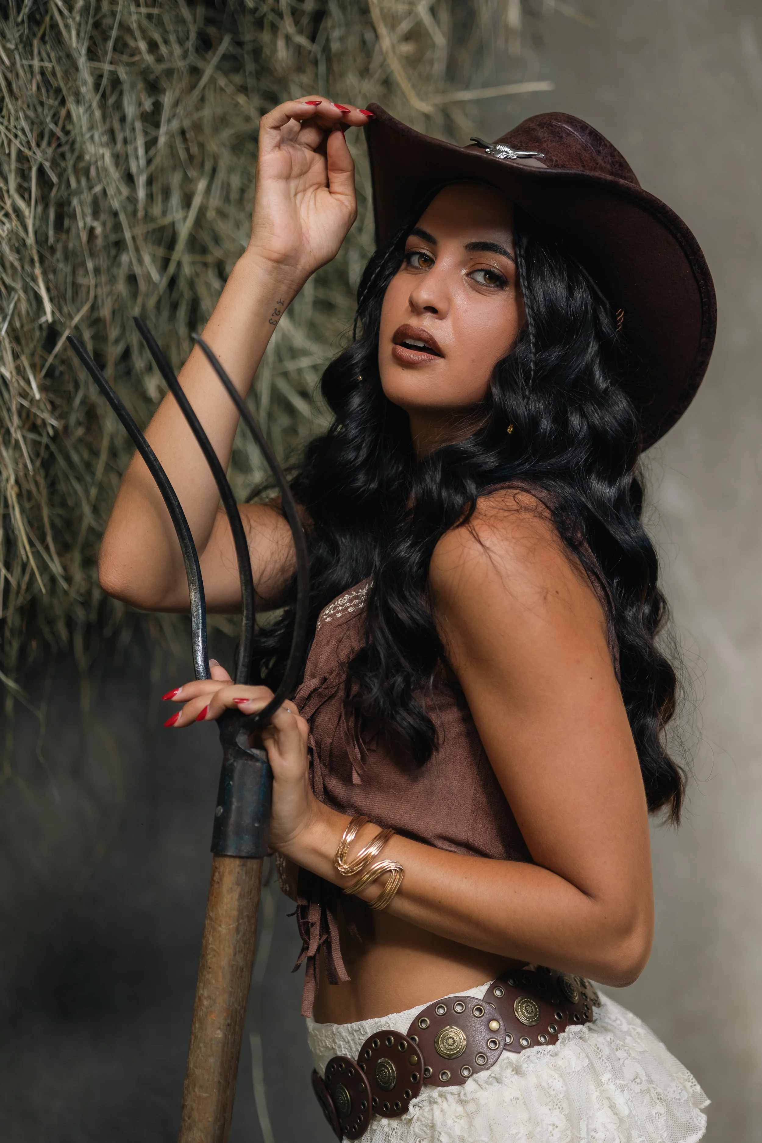 Une femme avec des cheveux longs et bouclés, portant un chapeau en cuir marron, une robe sans manches en tissu marron, un bracelet en or et une ceinture big arrondie en cuir marron. Elle tient une fourche mais elle regarde vers l'appareil photo avec