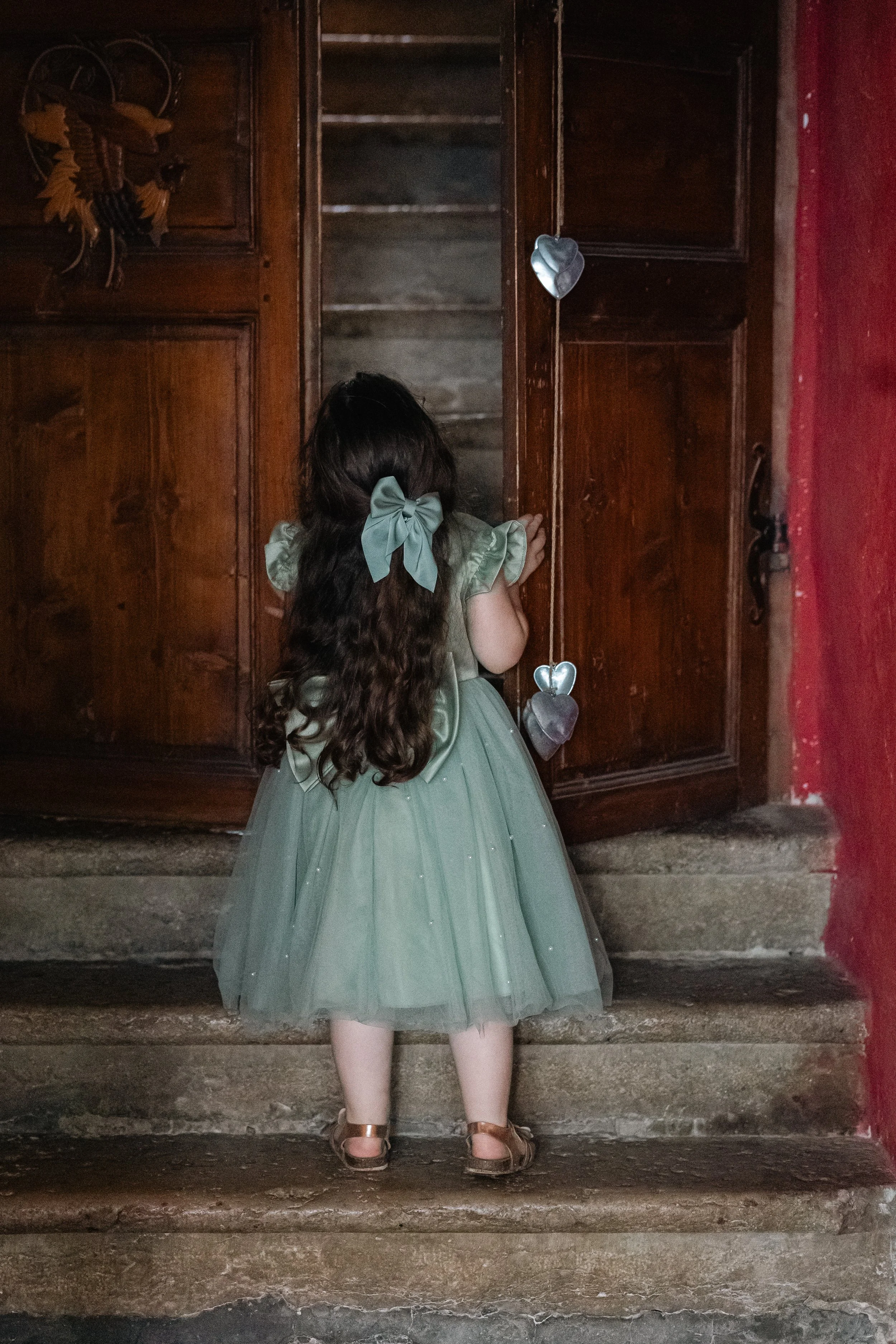 Une jeune fille portant une robe verte avec un gros nœud dans ses cheveux, regarde une porte en bois ancien dans une maison.