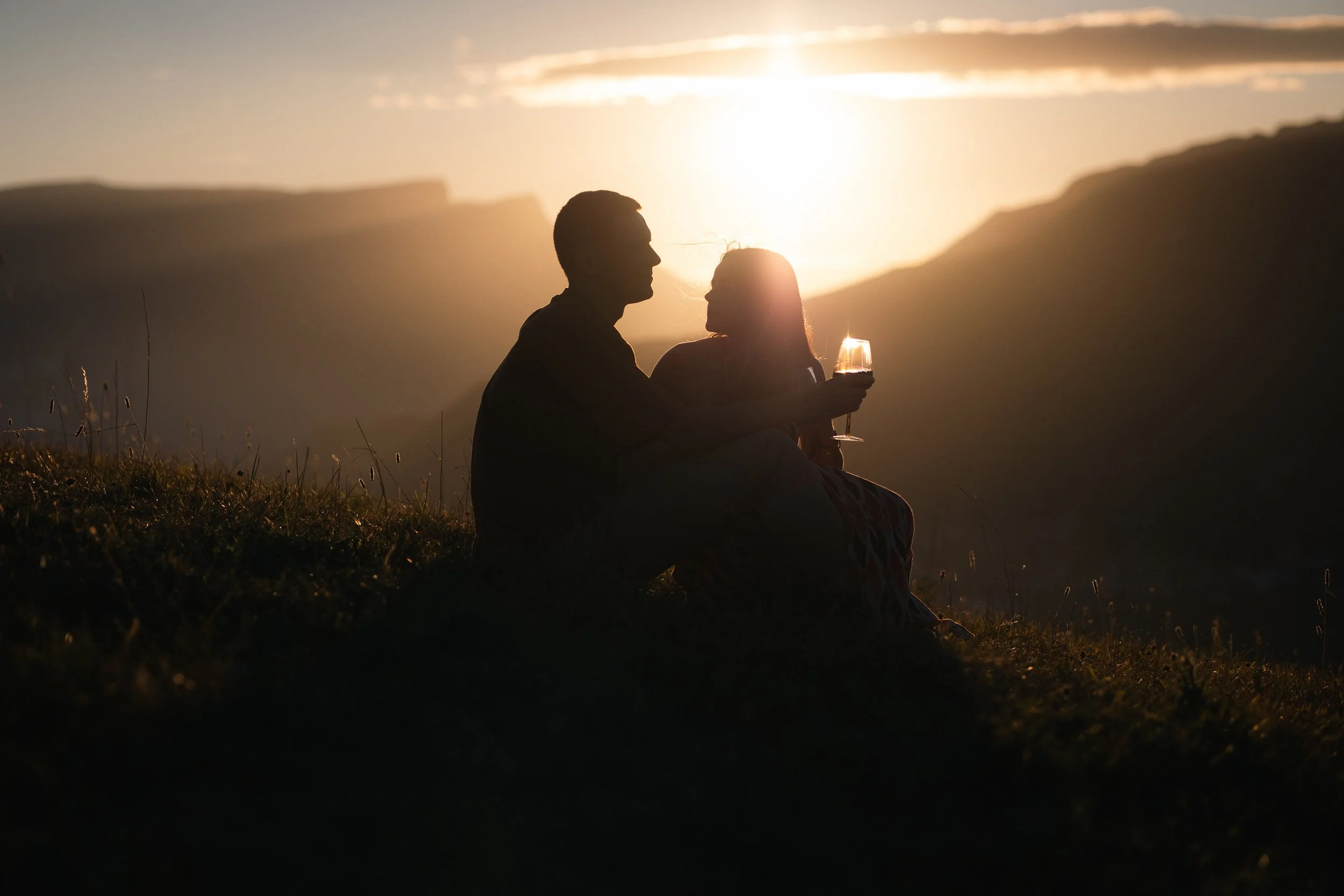 Un couple assis sur l'herbe lors du coucher de soleil, tenant un verre de vin, en silhouette contre le ciel lumineux.
