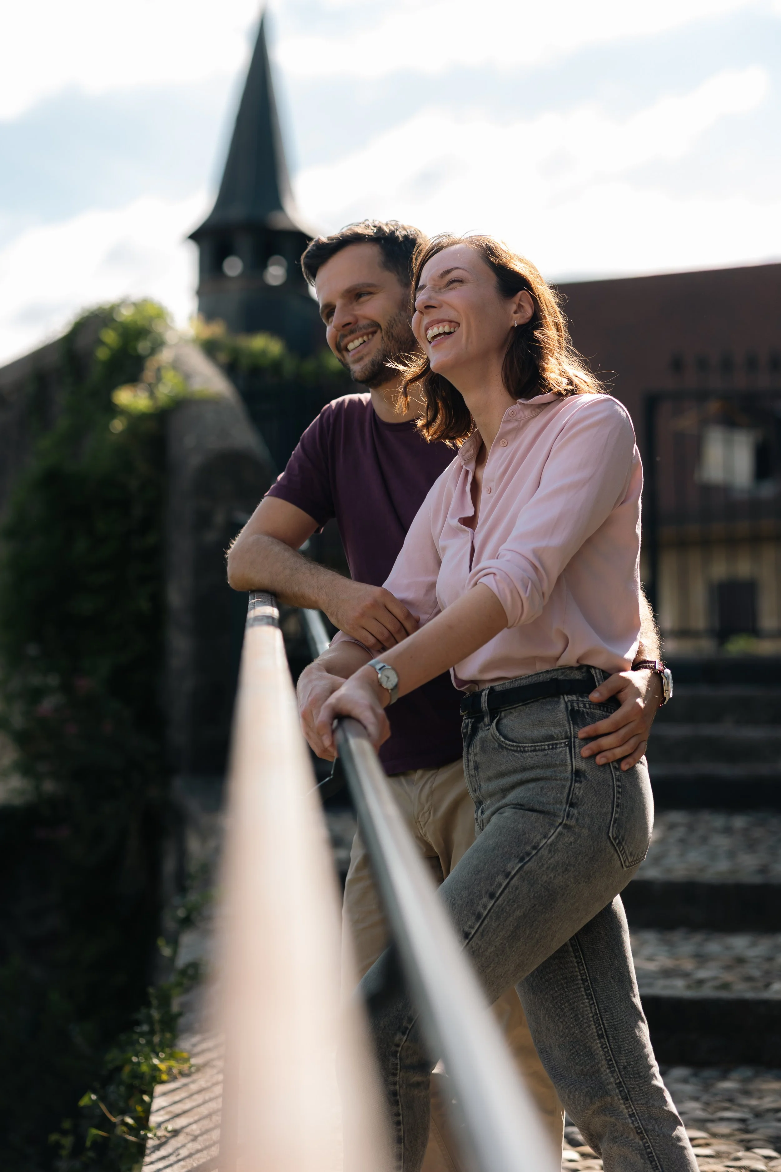 Un couple souriant, debout sur une rampe en extérieur avec un bâtiment avec un clocher en arrière-plan, sous un ciel partiellement nuageux.
