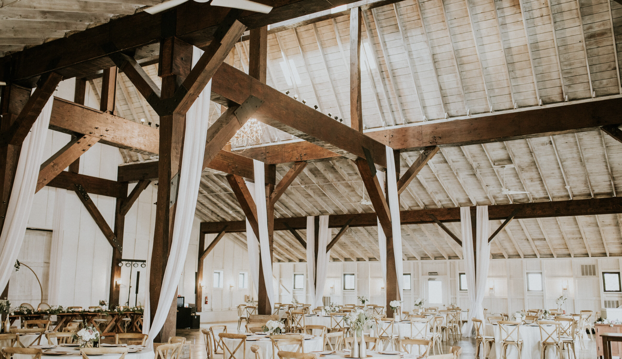 Interior of a rustic banquet hall with wooden beams, white drapery, and tables set with floral centerpieces and place settings for a wedding or event.