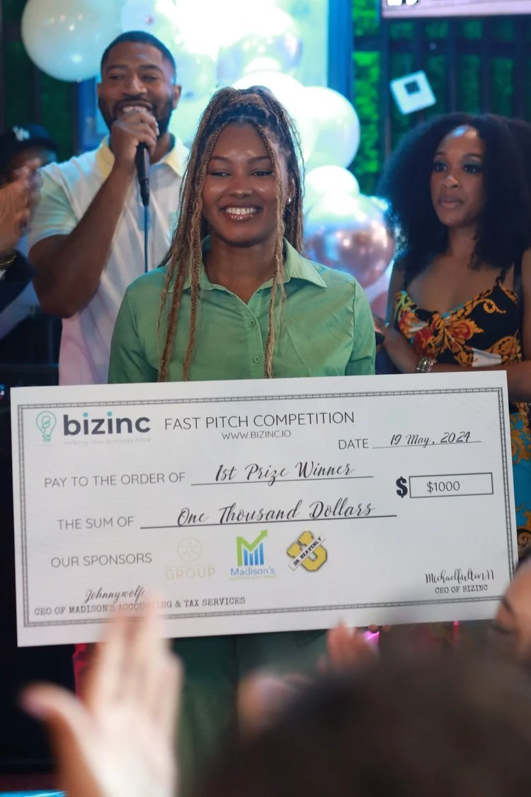 Smiling woman holding a large check for first prize at a pitch competition with people and balloons in the background.