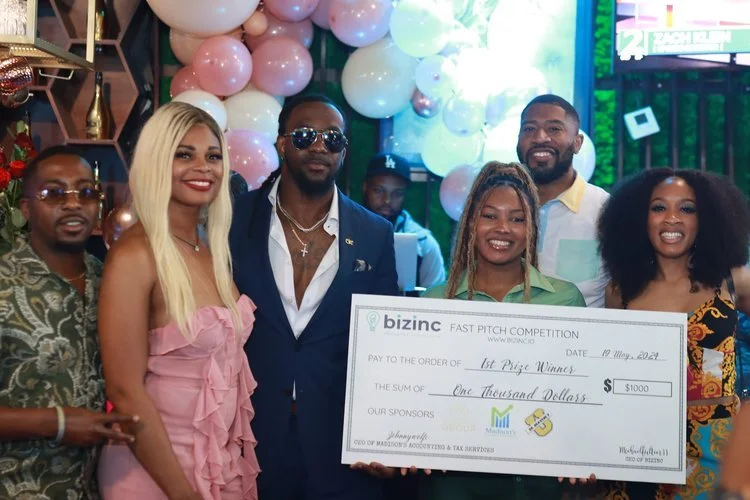 Group of six people posing with a large check for $1,000, indicating they are winners of a fast pitch competition, surrounded by balloons.