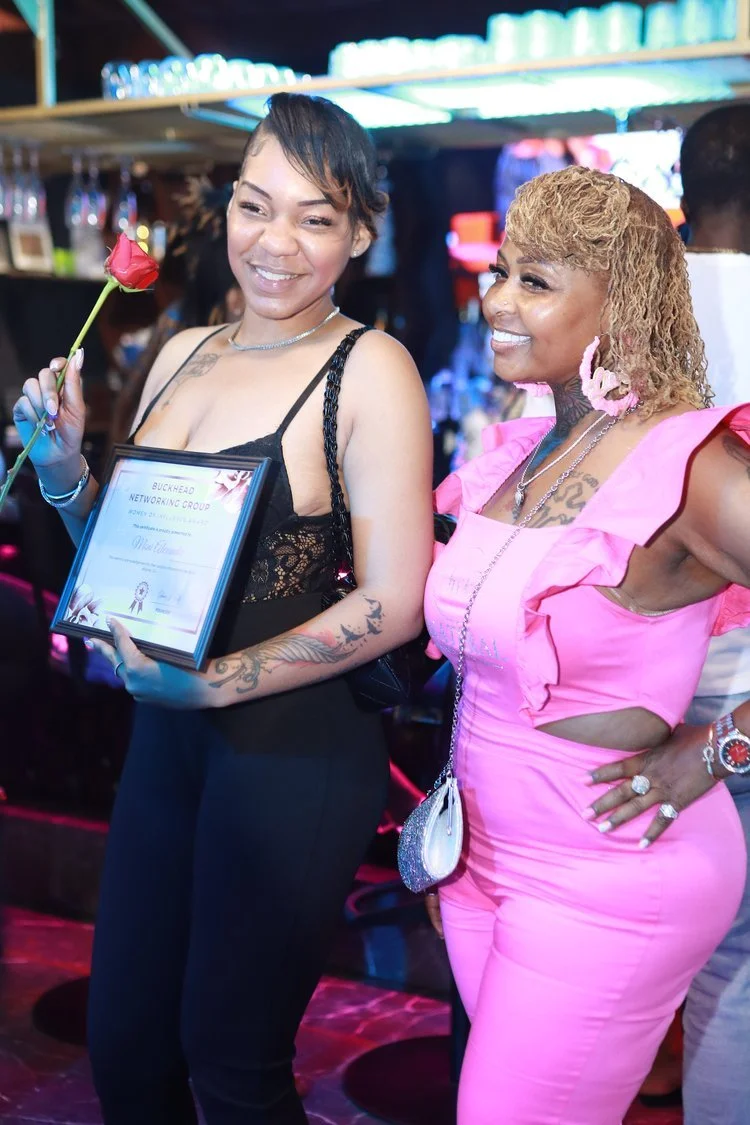 Two women posing at an event, one holding a rose and a certificate, the other in a pink dress. Background features shelves with glasses.