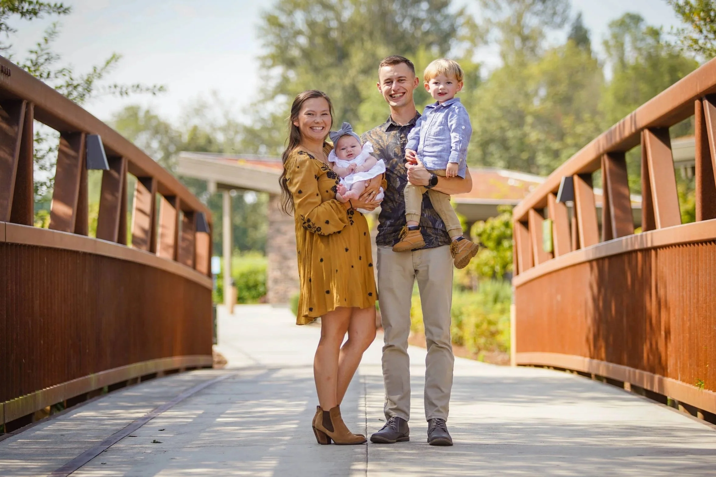 Family of four walking on a wooden bridge in a park on a sunny day, smiling and holding their young children.