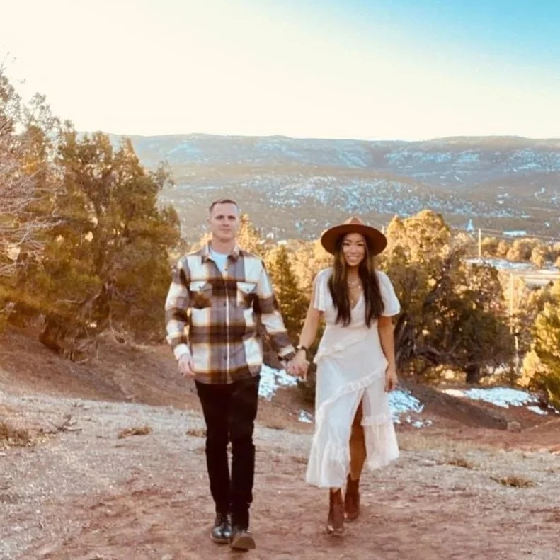 A couple walking hand in hand outdoors on a dirt trail in a hilly area with trees and snow patches, during sunset or sunrise.