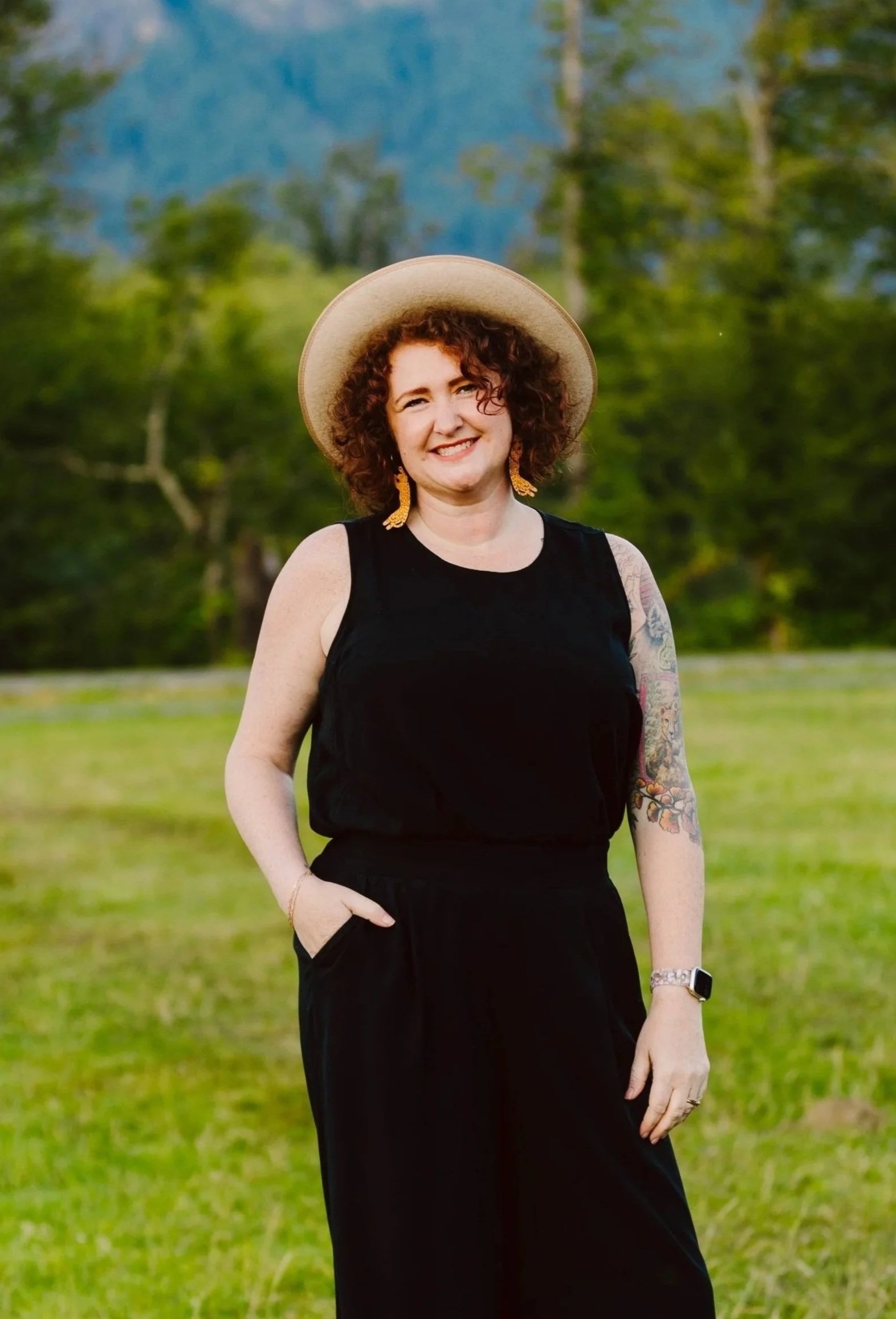 A woman with curly hair wearing a wide-brimmed hat and a black sleeveless dress, standing outdoors in a park or forest with mountains in the background.