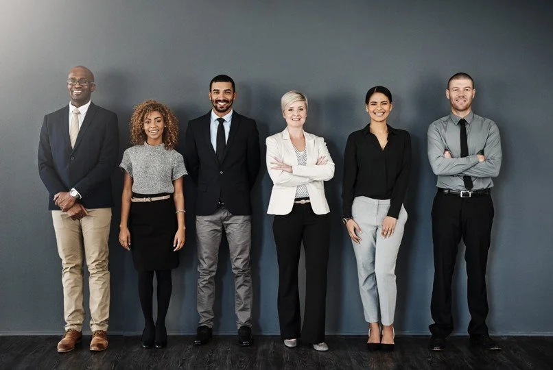 Group of six diverse professionals standing against a plain gray wall, dressed in business attire.