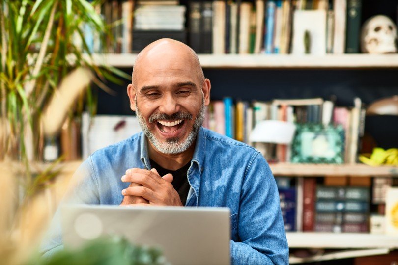 A smiling man with a beard and bald head sitting at a table with a laptop in front of him, in a room with a bookshelf and decorative items in the background.