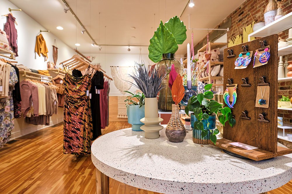 Inside a boutique store with clothing on racks and a central table displays decorative plants, vases, and jewelry on a wooden stand.