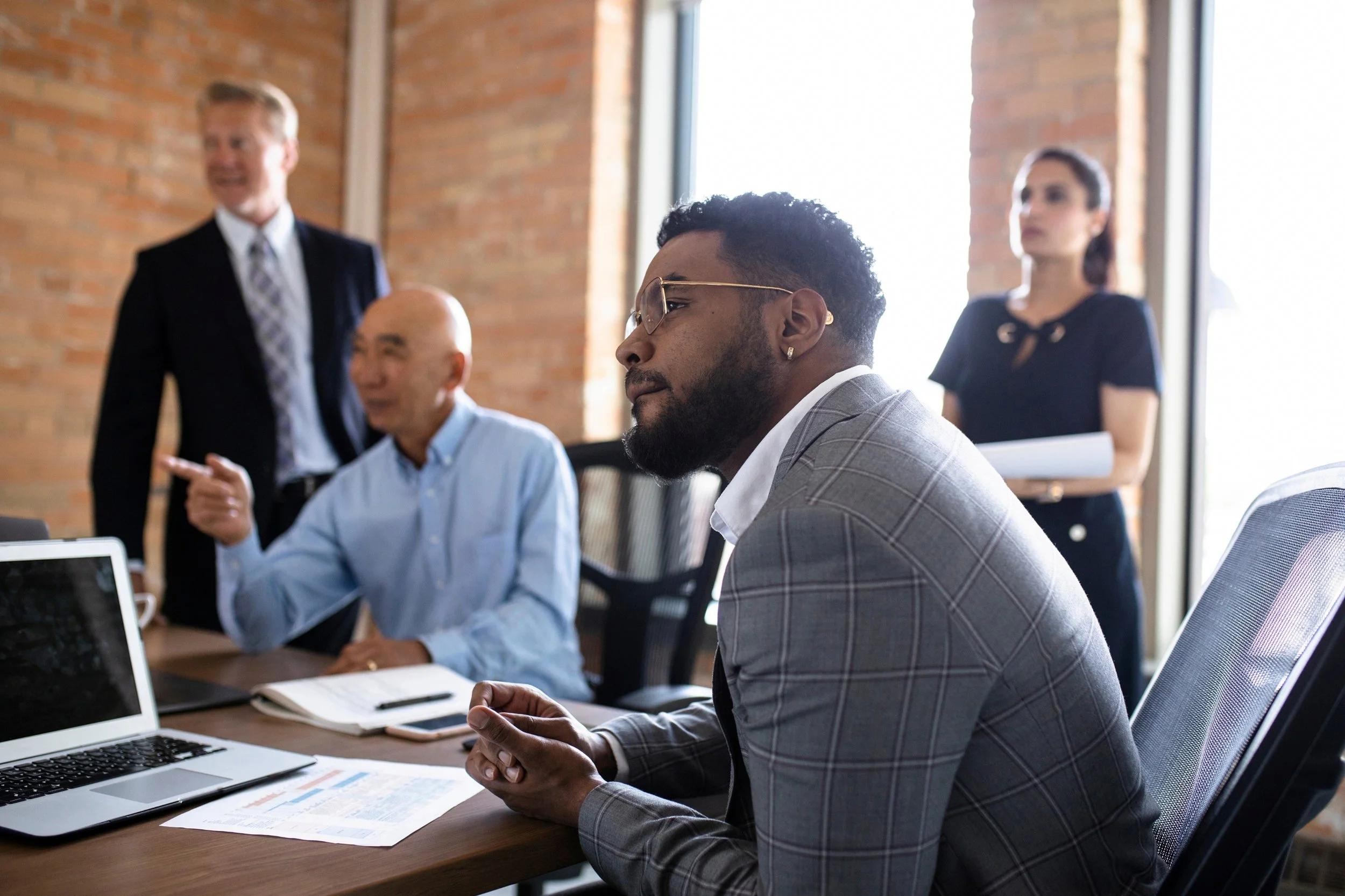 Business meeting with four people in a modern office, with exposed brick walls and large windows, one man is sitting at a desk, looking thoughtful, two people are standing in conversation, and a woman is standing with a notepad.