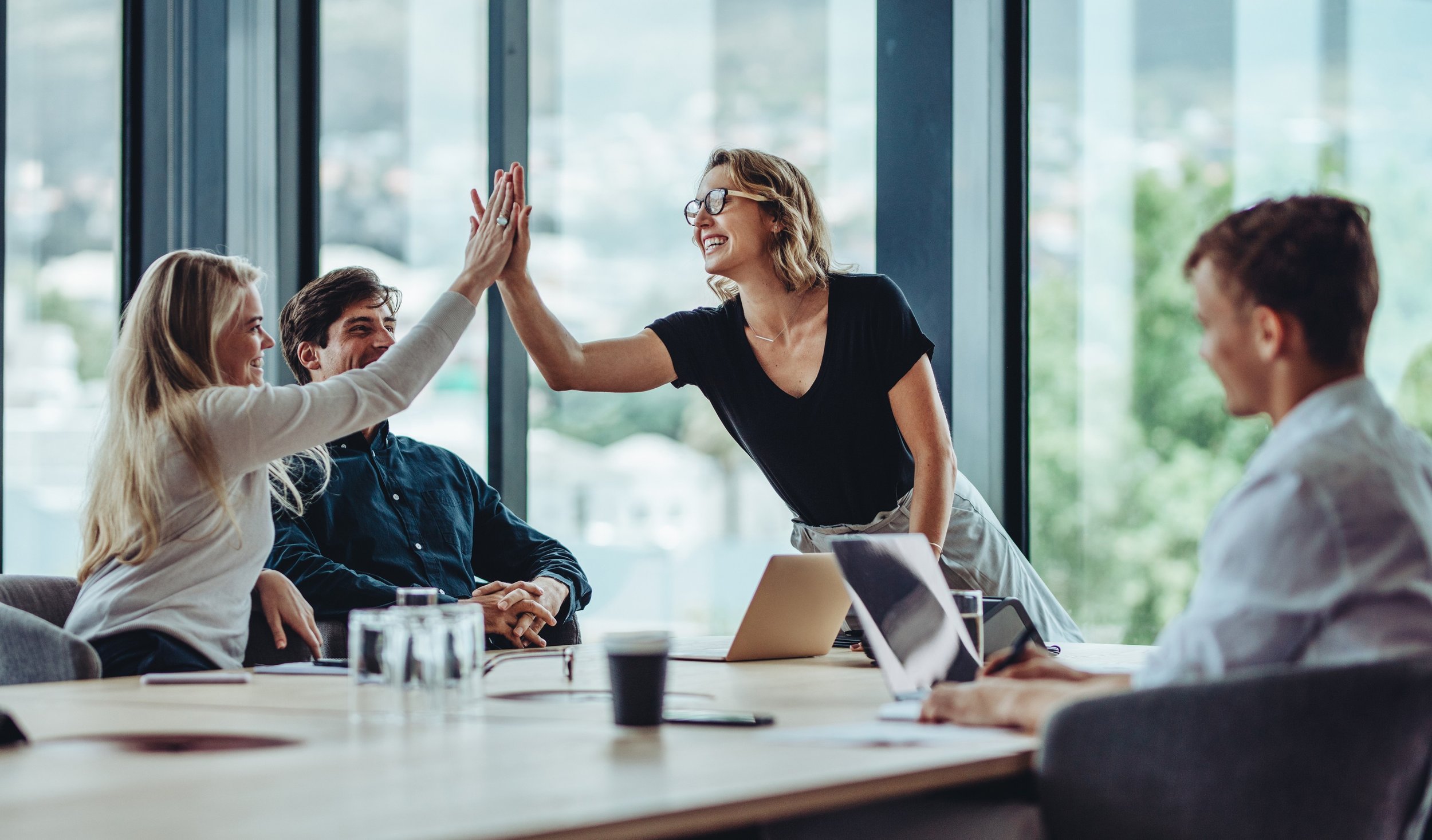 Group of four coworkers in a modern office celebrating with high fives around a conference table.
