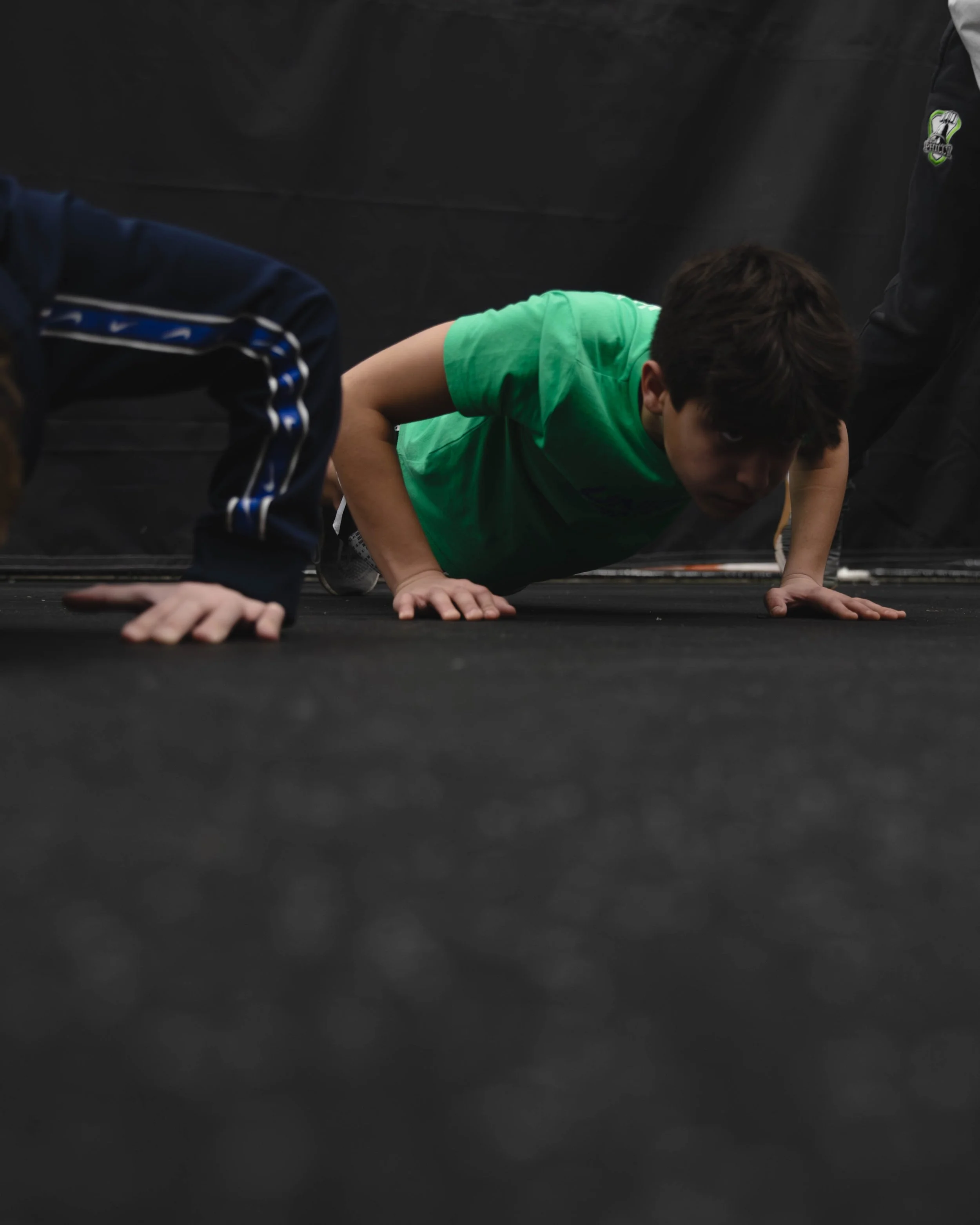 Youth performing push-up exercise on black gym floor, wearing green shirt, dark hair, and dark background.