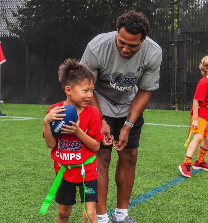 A young boy in a red t-shirt holding a blue football on a football field, smiling as he talks to an adult man who is leaning down and smiling back. The boy is wearing a green flag around his waist. In the background, another child in red and orange athletic clothing is visible.