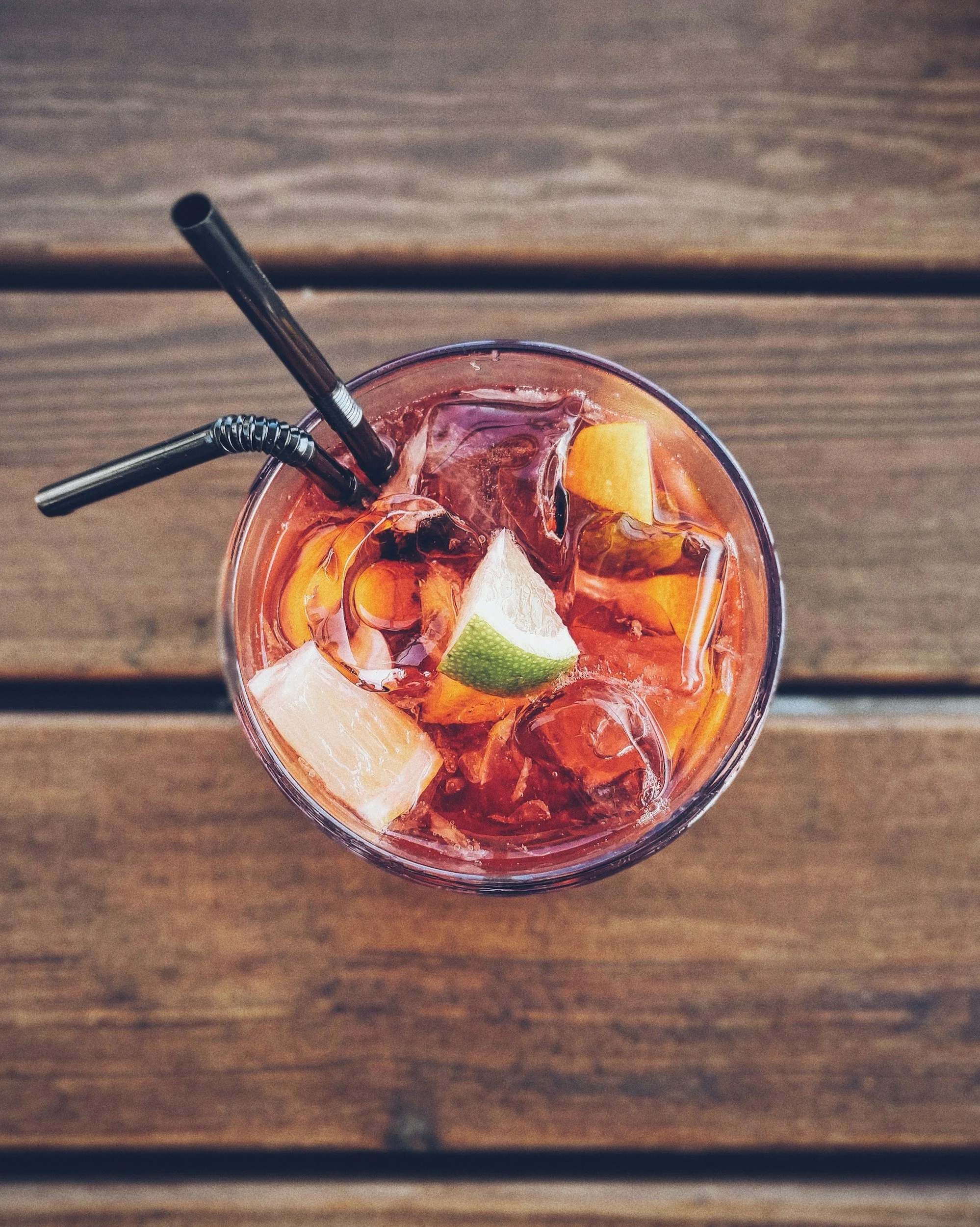 Top view of a glass of iced drink with orange, lime, and lemon slices, with two black metal bendable straws, on a wooden surface.