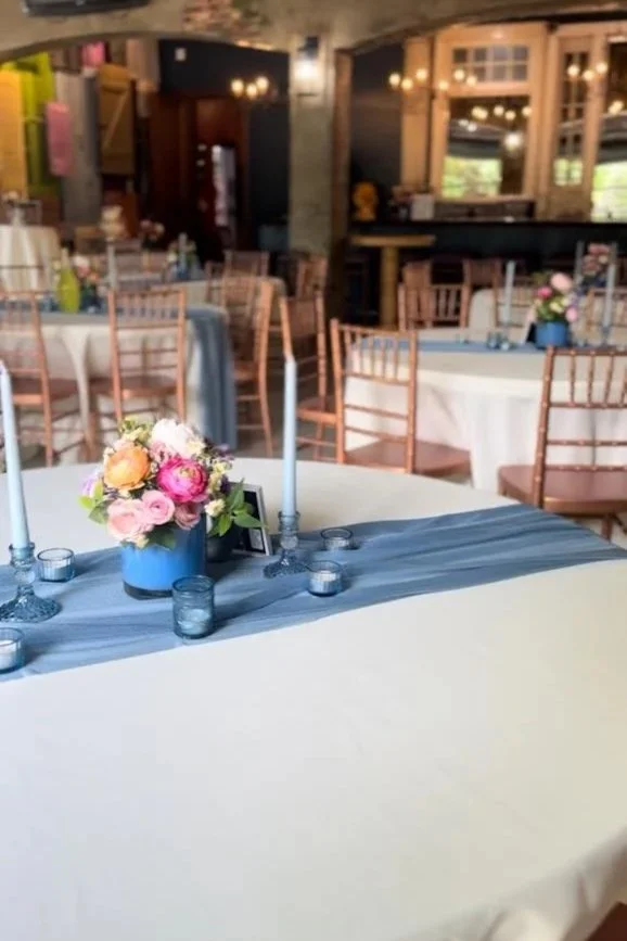 A round banquet table decorated with a blue flower arrangement, blue candles, and small votive candles in a rustic, well-lit event space with wooden and stone decor.