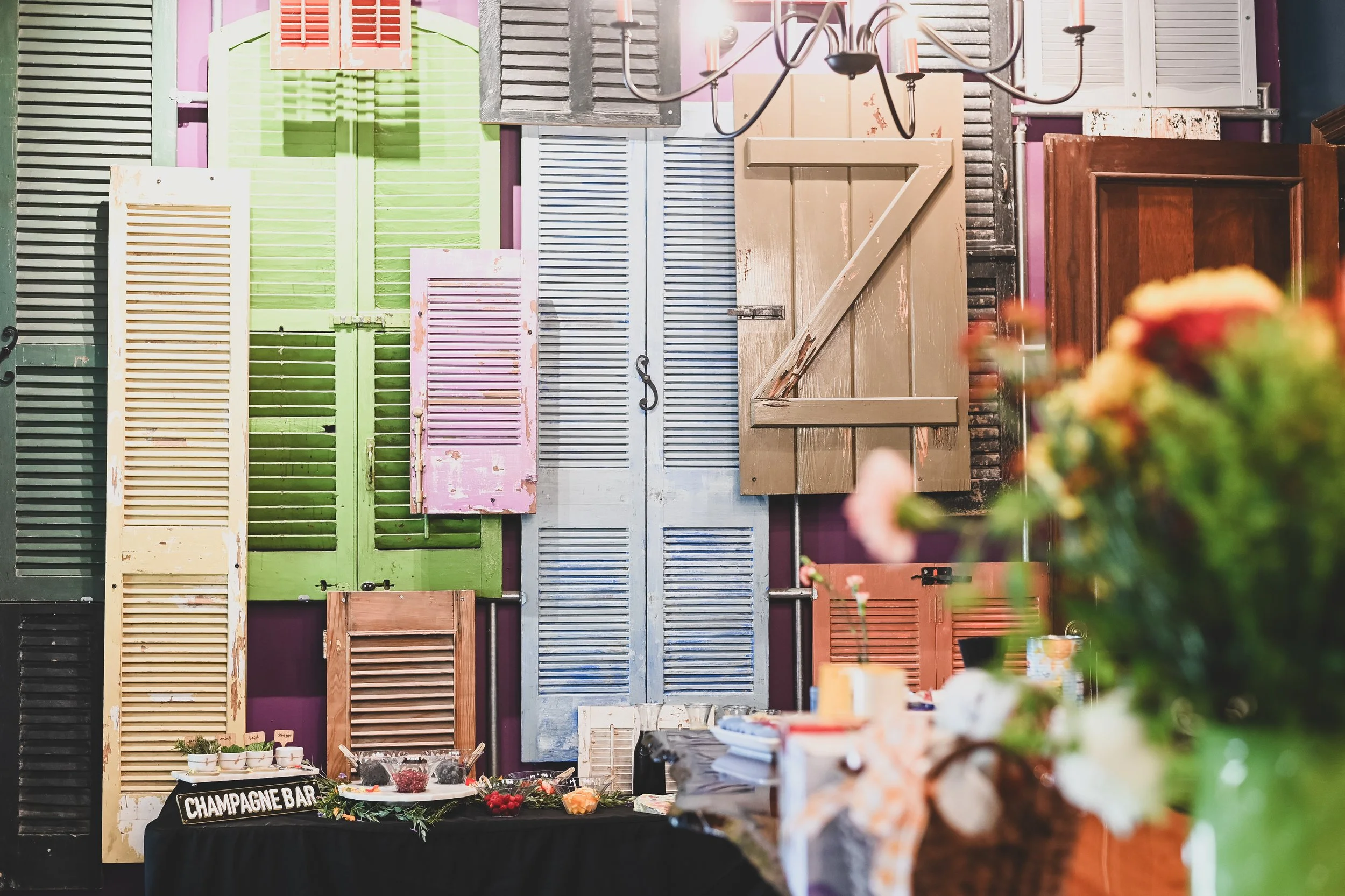 A display of colorful vintage shutters on a purple wall, with a chandelier hanging above. In the foreground, a table with a sign that reads 'Champagne Bar' is decorated with flowers and bowls of berries and other small food items.