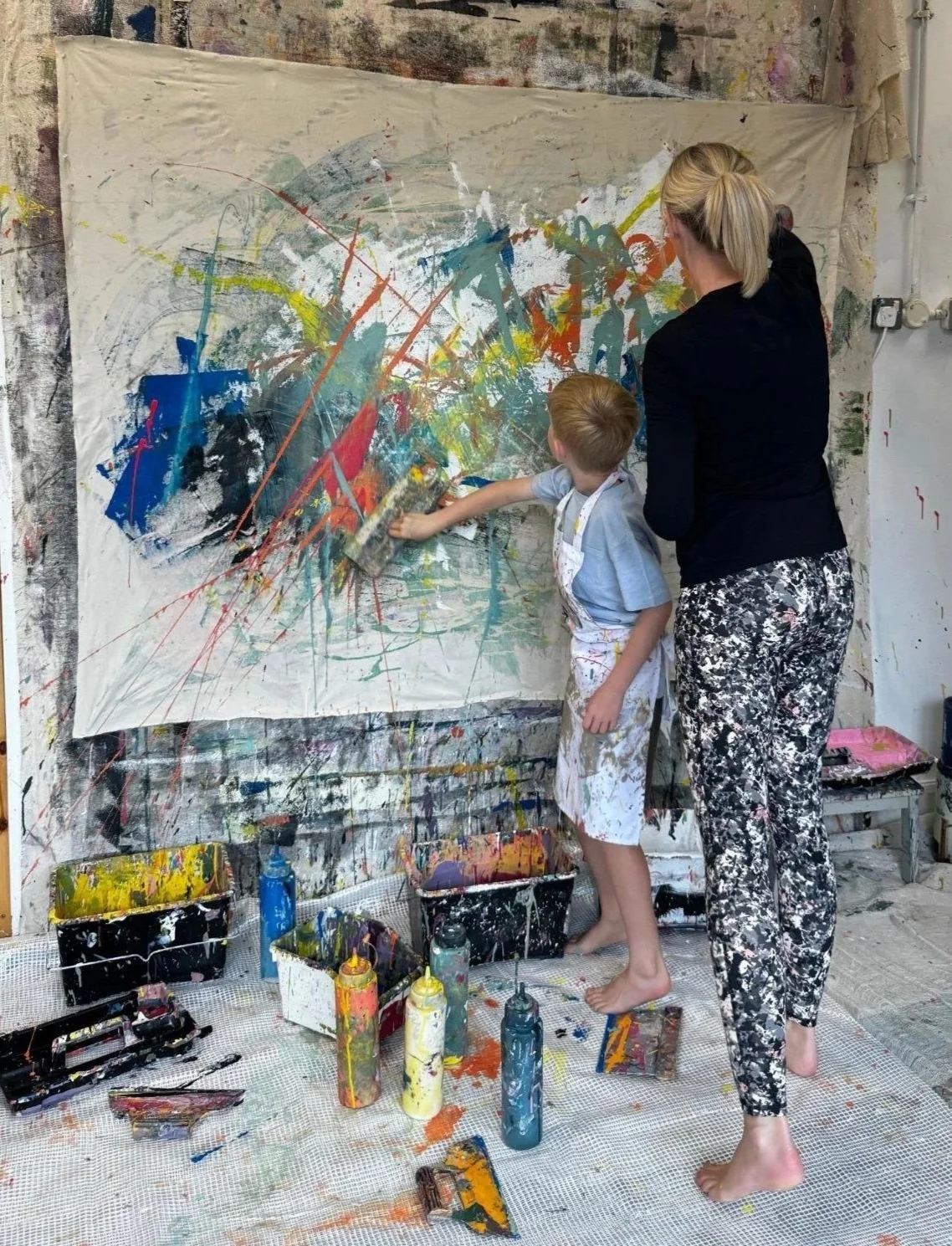 A woman and young boy creating an abstract painting on a large canvas in an art studio, with paint bottles, brushes, and splatters surrounding them.