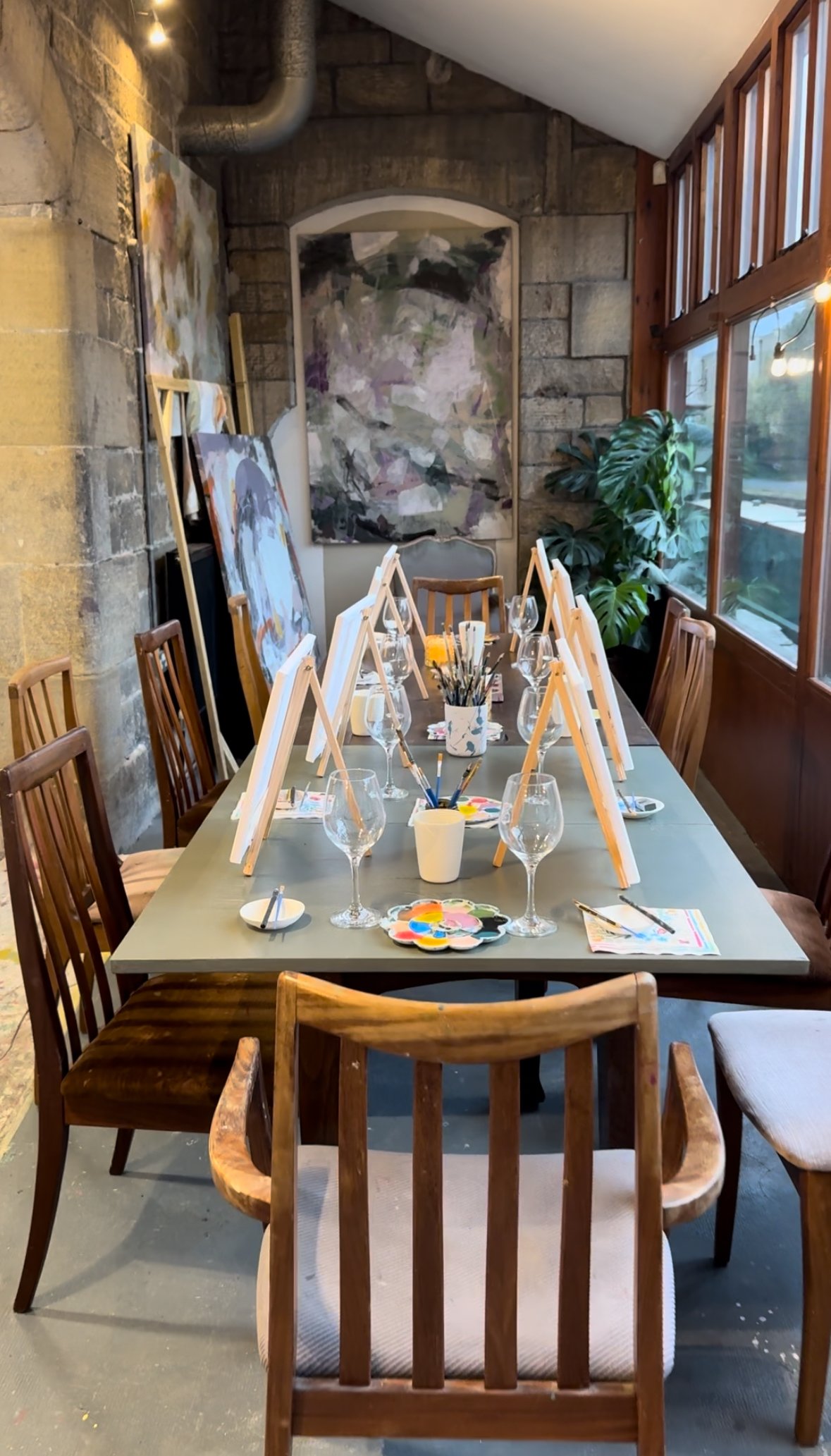 A dining table set up for painting with easels, brushes, paints, wine glasses, and a centerpiece, in a cozy room with stone and wood walls, large windows, and artwork.