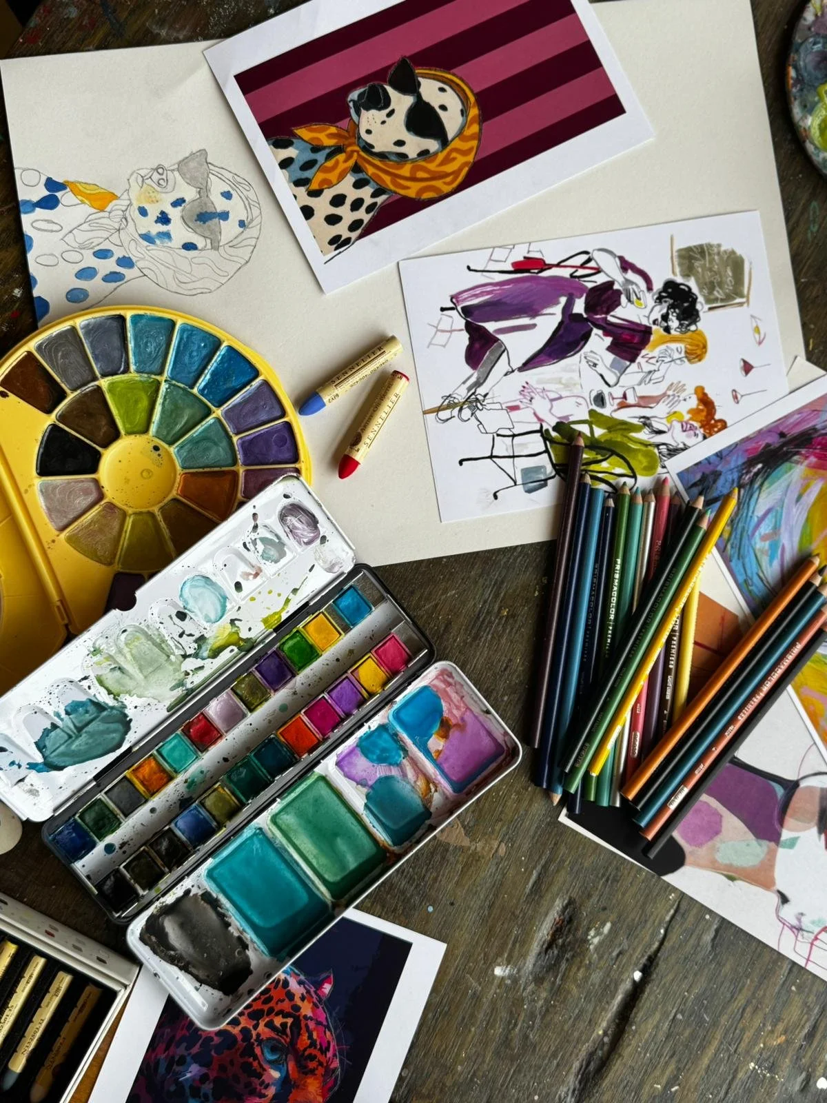 A messy artist's workspace with watercolor paints, colored pencils, sketches, and finished paintings, including dog and leopard artworks.