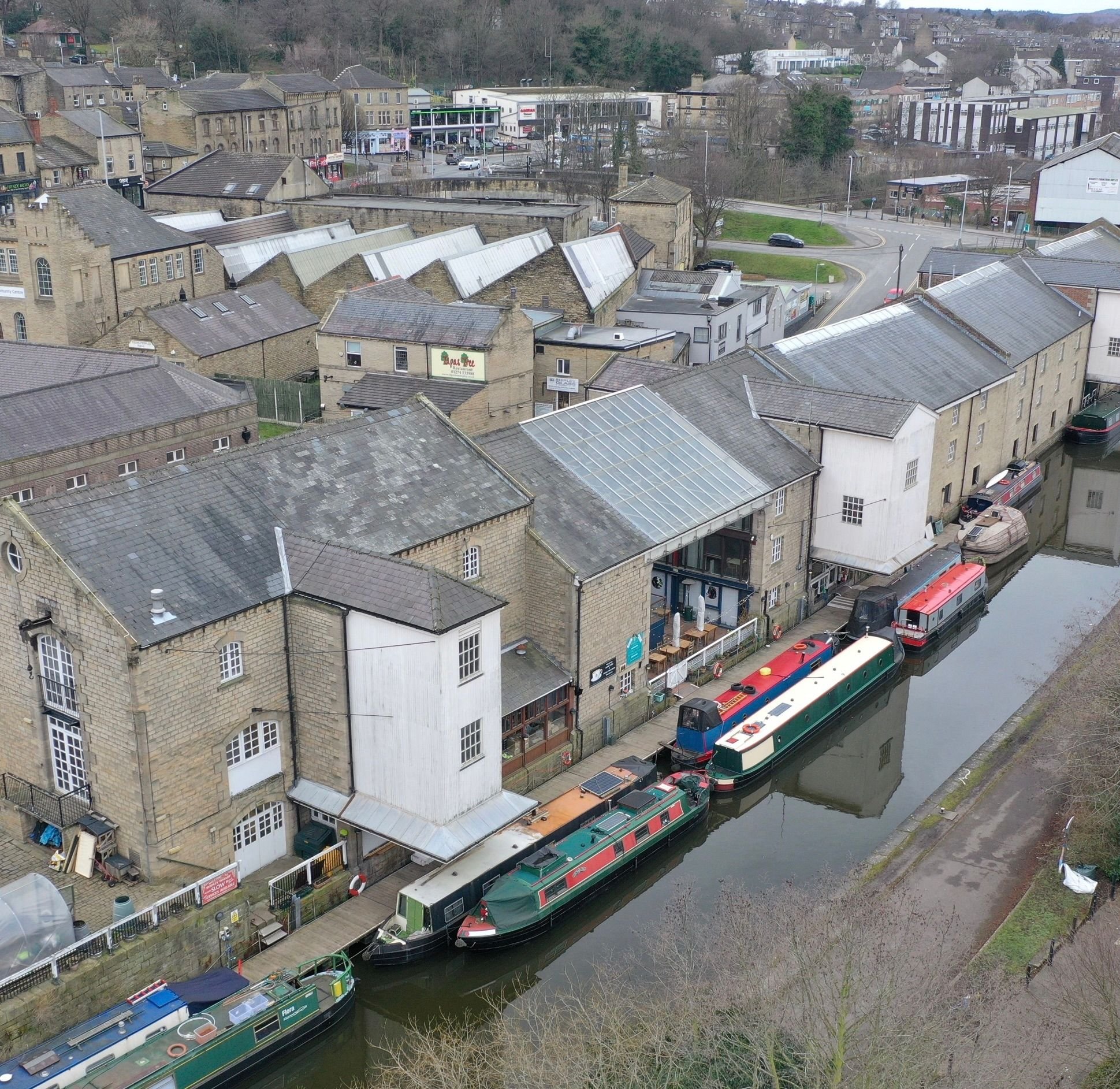 Aerial view of a canal with narrowboats in a town, surrounded by old brick buildings, some with rooftop glass structures, and streets with cars in the background.