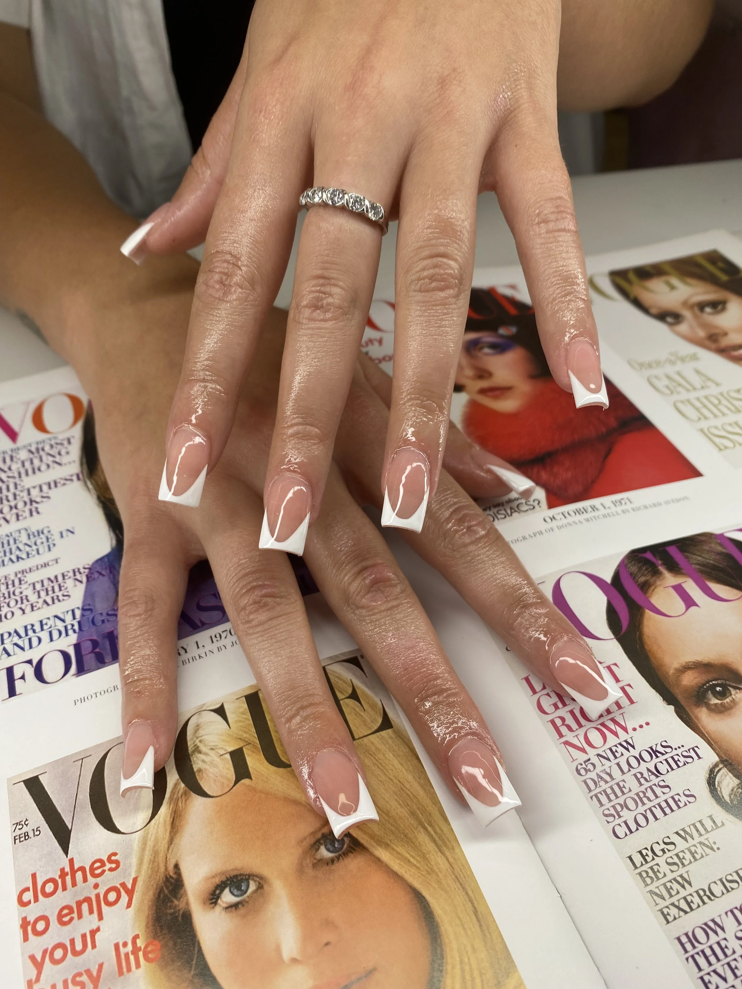 A woman's hands with freshly manicured nails featuring white French tips, resting on Vogue magazine covers. She wears a silver ring with multiple stones on her finger.