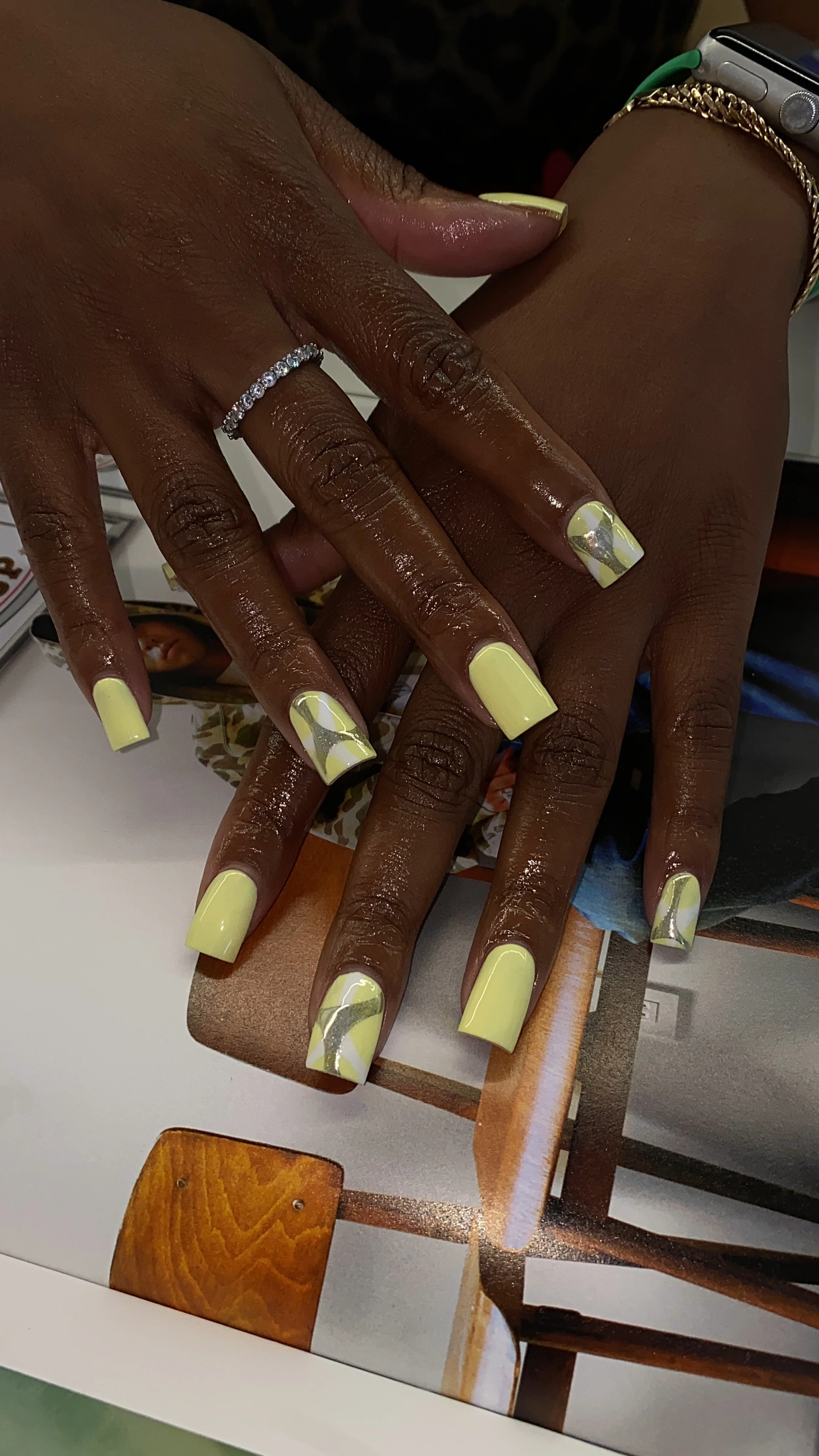 Close-up of a woman's hands with fresh yellow and silver marbled nail polish on her nails, wearing rings and bracelets, resting on a magazine or book with wooden chair and furniture in the background.