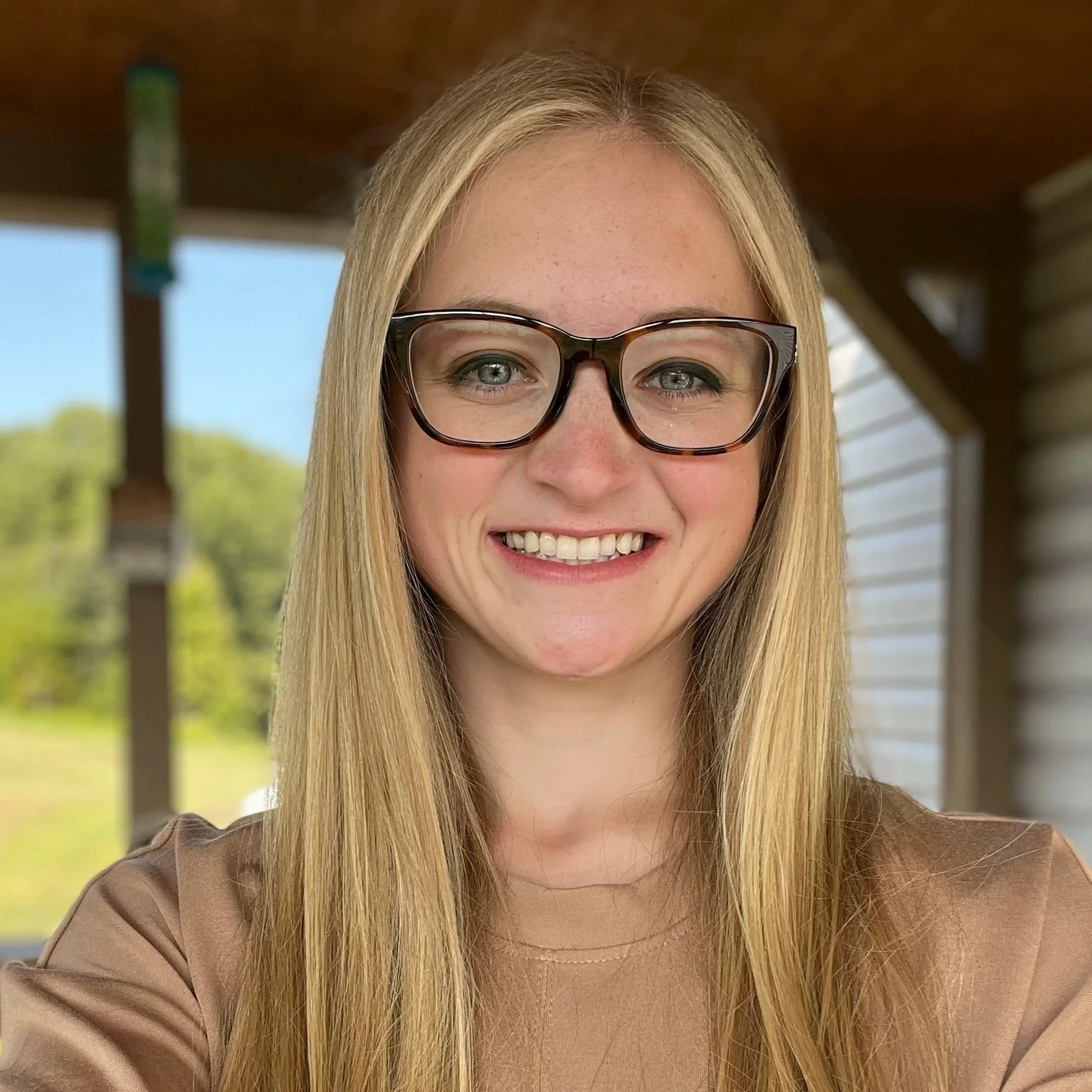 Close-up selfie of artist Emerald Downey with blonde hair, glasses, and a friendly smile, outdoors under a structure with a wooden roof and blurred greenery in the background.
