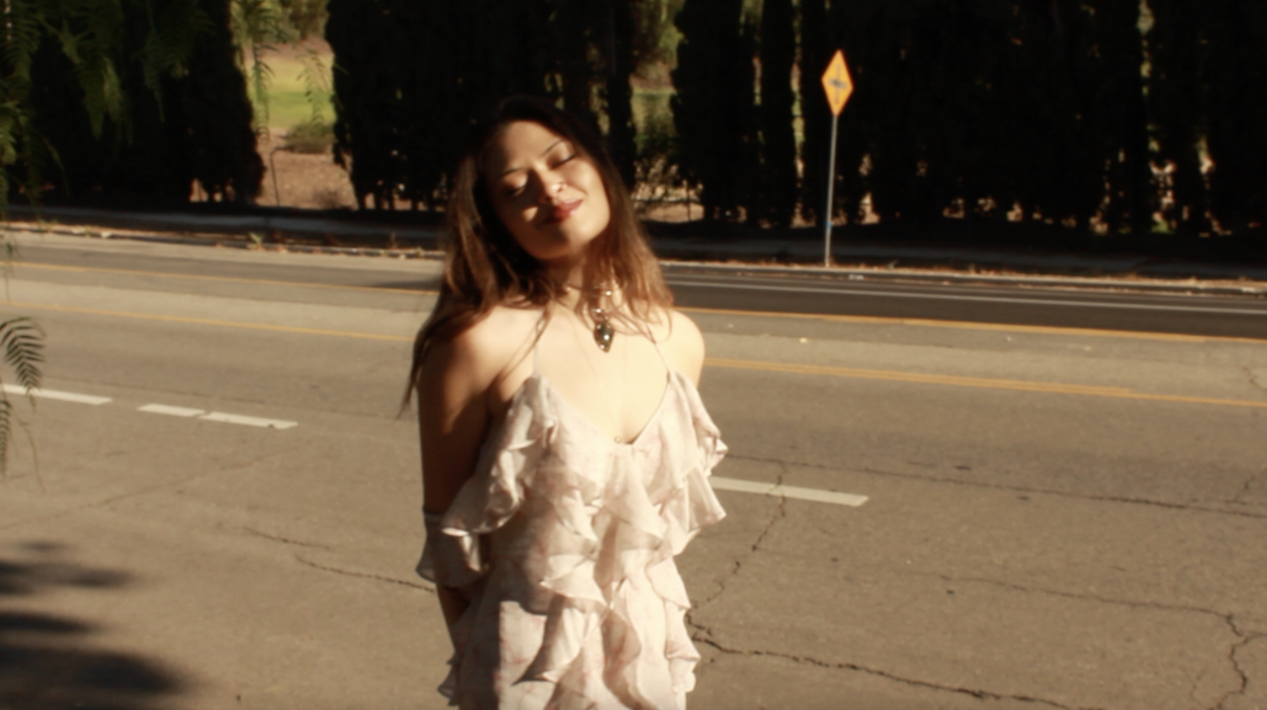 A young woman with long brown hair is standing on the side of a road with trees in the background. She is wearing a light-colored, ruffled dress and jewelry, smiling with her eyes closed.