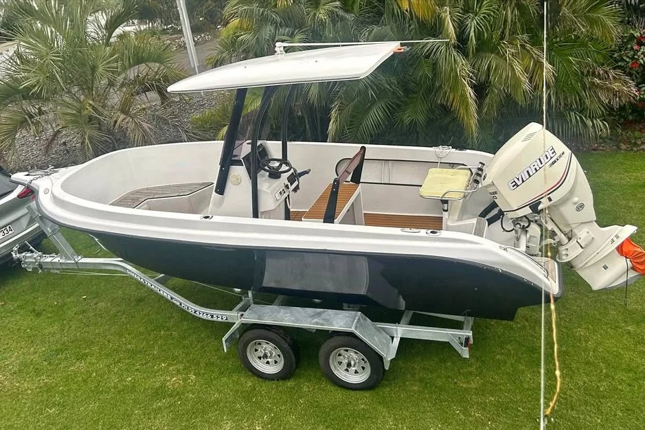 A small boat with a black hull on a boat trailer, equipped with a white outboard motor, parked on a grassy area with palm trees in the background.