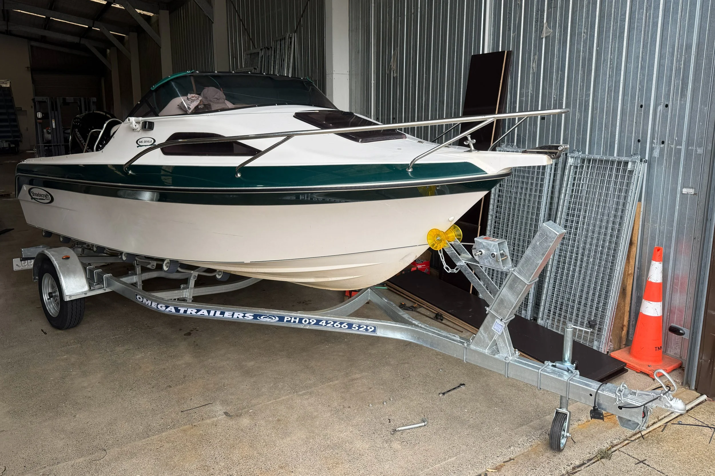 A small white and green motorboat on a trailer inside a metal garage, with orange traffic cone and metal grates against the wall.
