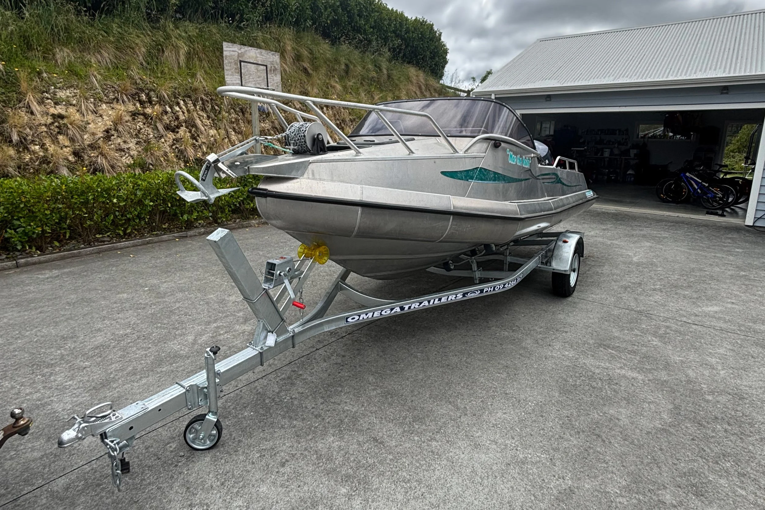 A small motorboat on a boat trailer parked on concrete in front of a garage, with grass and hills in the background.
