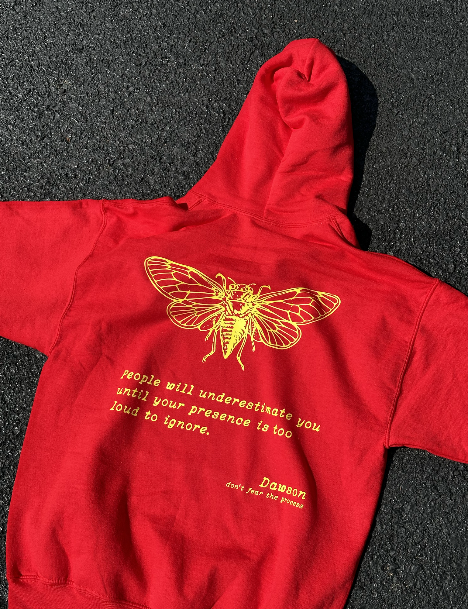 Red hoodie with a gold cicada graphic and the quote "People will underestimate you until your presence is too loud to ignore"