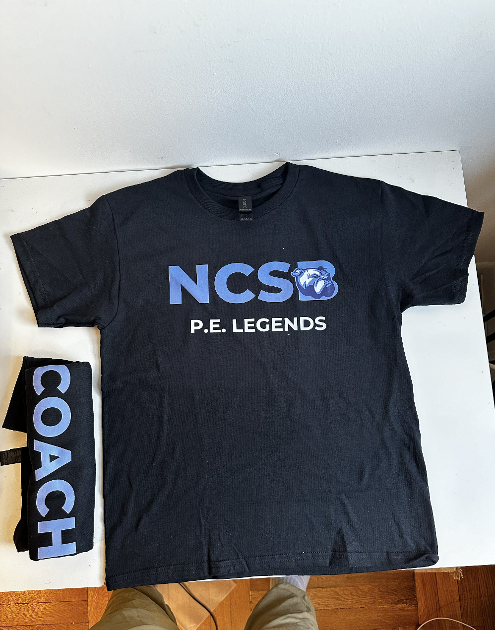 Black "NCS P.E. Legends" tee with a "Coach" sleeve print