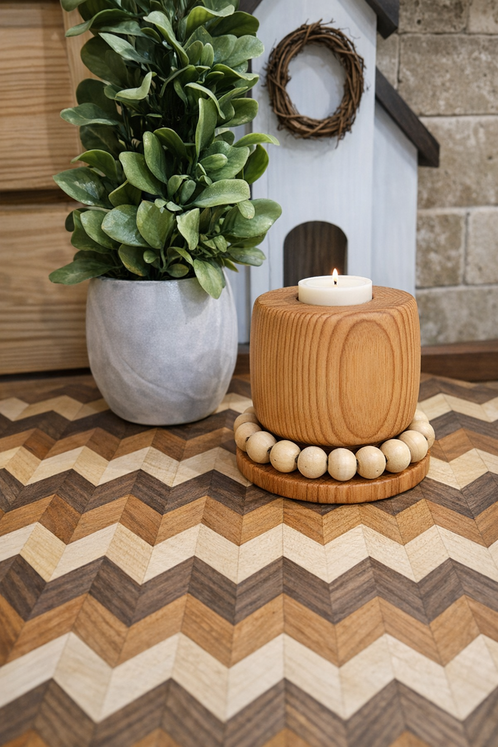 Decorative interior scene featuring a potted green plant, a wooden candle holder with a lit white candle, and a small white house-shaped decor with a wreath, all arranged on a patterned wooden surface.