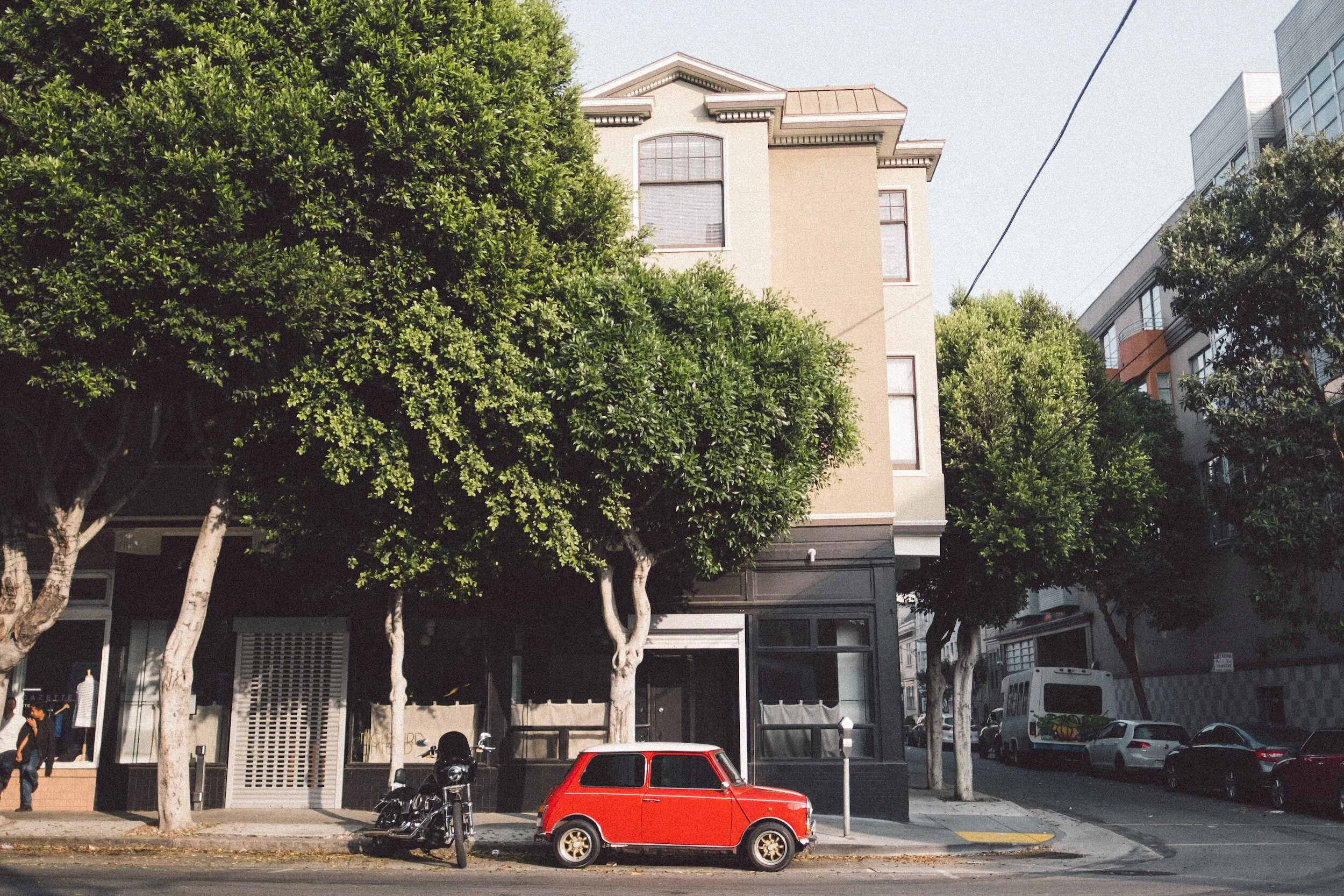 Street scene with a small red car and motorcycle parked near a beige building with large trees.