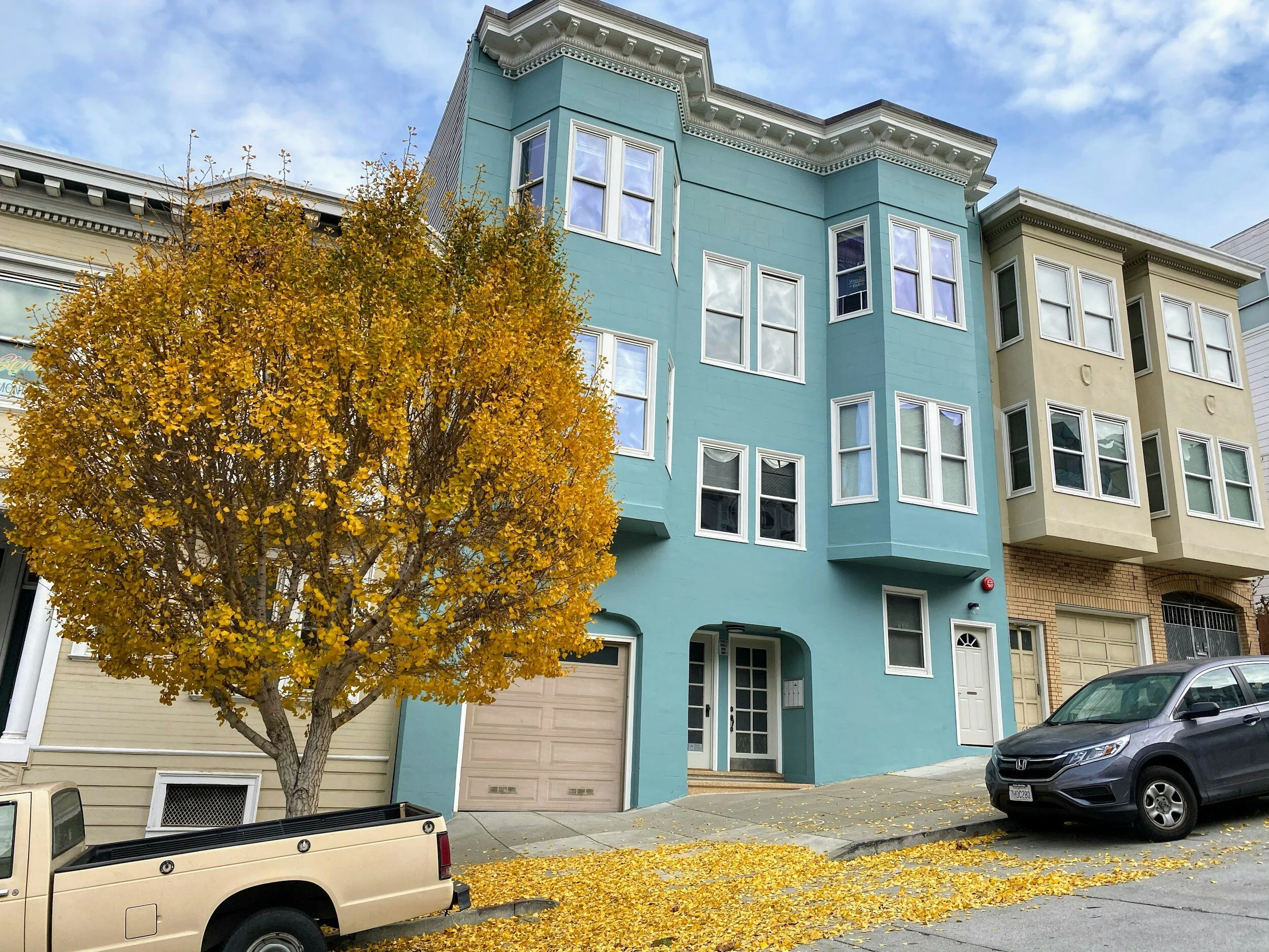 Row of three-story houses in a residential neighborhood, featuring a blue building with bay windows. A large tree with yellow autumn leaves stands on the sidewalk, near a beige pickup truck and a gray car.