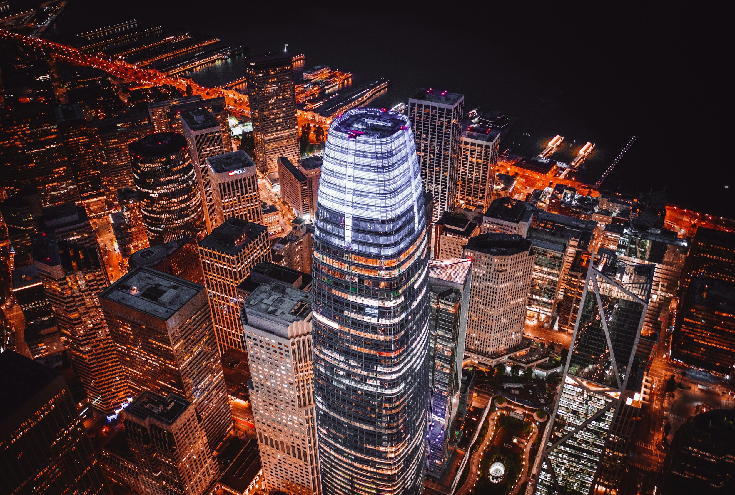 Aerial night view of a lit skyscraper in a city, surrounded by other illuminated buildings and streets with light trails.