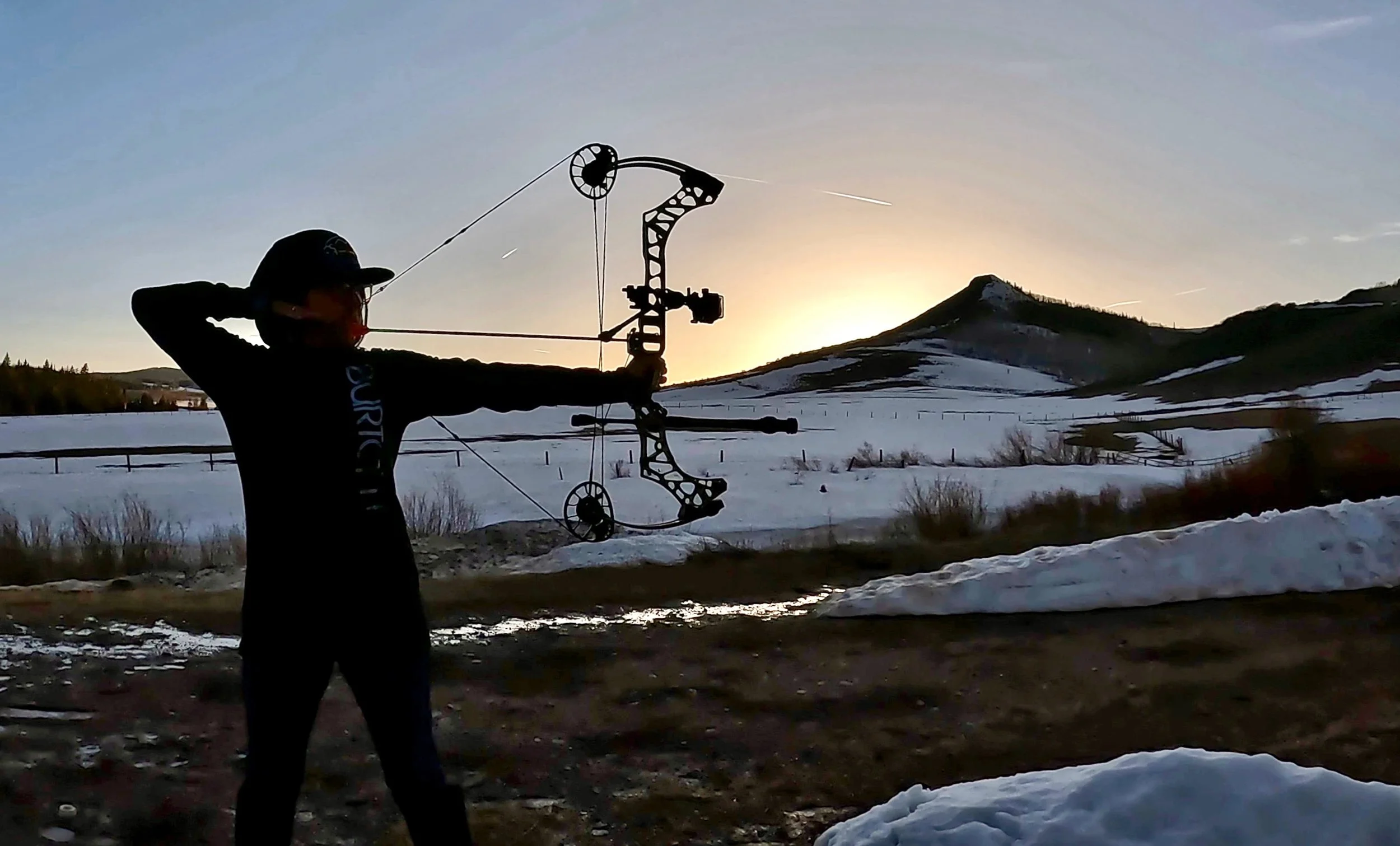 Person shooting a bow outdoors during sunset in a snowy landscape with mountains in the background.